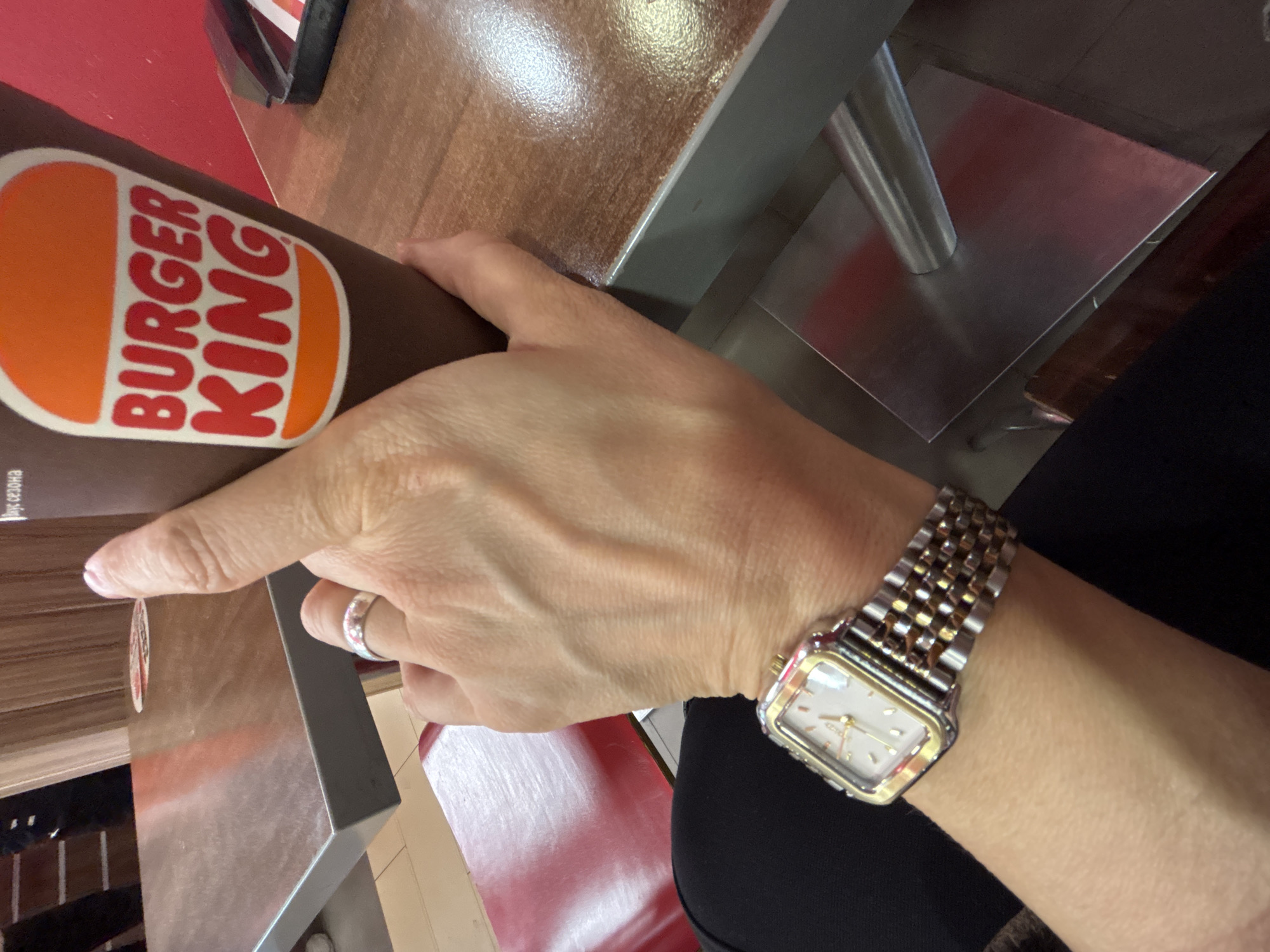The image shows a person's hand wearing a watch with a metal band and a silver ring on the ring finger. The hand is holding a Burger King container. The scene appears to be inside a Burger King restaurant, evidenced by the container and the table design. The image does not show any elements to determine the exact location within Izhevsk, Russia.