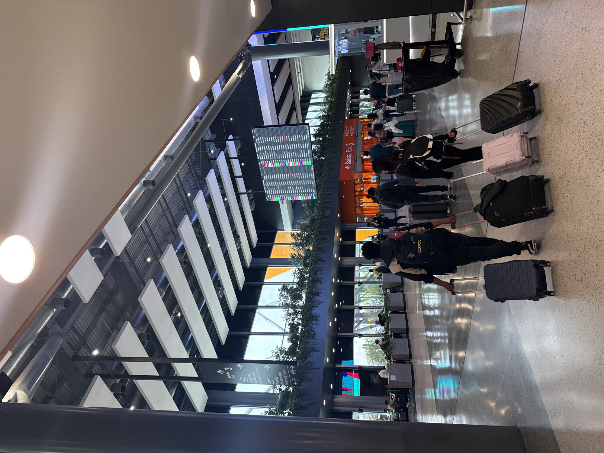 The image depicts a bustling indoor airport terminal, likely a departure or waiting area, located in Apodaca, Mexico. The scene is dominated by numerous travelers, mostly seen from behind, standing in queues or moving through the space, accompanied by various pieces of luggage. Suitcases of different sizes and colors, including black, pink, and dark blue, are visible on the polished, light-colored floor.
The terminal features modern architecture with high ceilings, supported by robust pillars, and illuminated by a combination of natural light filtering through large windows and numerous recessed artificial lights. Overhead, decorative white rectangular panels and exposed ventilation ducts contribute to the industrial yet contemporary aesthetic. Rows of airport seating are visible in the background, where some people are resting.
A large digital flight information display screen is centrally located, showing a grid of flight details, including flight numbers, destinations, times, and statuses, indicated by various colored segments (red, green, blue). Prominent directional signage is visible, clearly indicating "Salida / Exit 3," "Llegadas / Domestic Arrivals," and "Puertas de Abordaje / Boarding Gates," providing clear context for an airport environment. Another "Salida / Exit 3" sign is visible further back. One person's backpack has the text "CCKPD" on it.
Lush greenery in planters lines some dividers, adding a touch of nature to the modern space. The overall activity indicates people engaged in the typical processes of air travel, such as waiting for check-in, security, or boarding. The lighting suggests it is daytime.