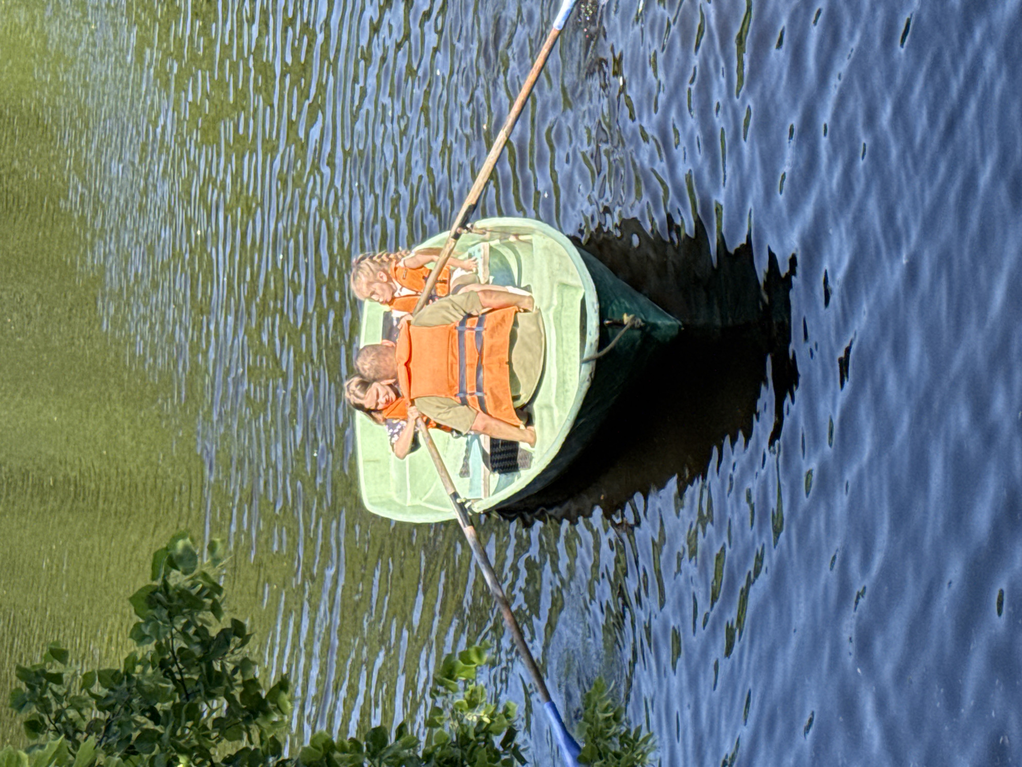 A light green rowboat with three occupants is on a body of water. In the center, a person is seated and rowing the boat with two wooden oars. In front of the rower, two children wearing orange life vests are seated. The child on the left is looking towards the rower, while the child on the right is looking out towards the water. The boat is surrounded by dark blue water with ripples, and green foliage is visible on the left side of the image. The water appears to be a natural body of water, possibly a pond or a small lake, within a park or natural area.