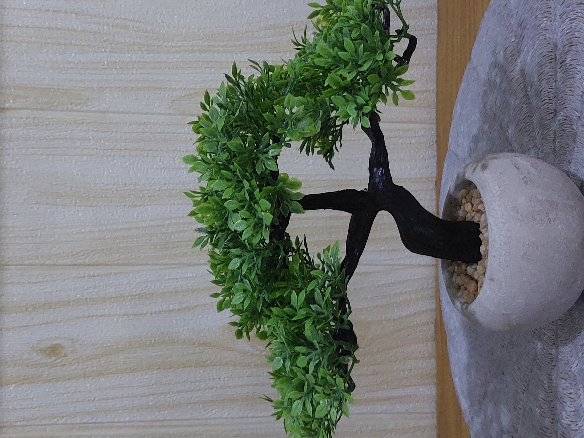 This is a close-up, eye-level, indoor shot of a small, artificial bonsai tree. The tree has a dark, twisted trunk and branches, adorned with numerous small, bright green leaves. It is planted in a light-colored, round pot filled with small, light brown pebbles. The pot rests on a textured, light gray surface, which in turn sits on a wooden table. Behind the bonsai tree is a wall with a light beige, vertical wood grain pattern. The image was taken in the City of Pematang Siantar, Indonesia.