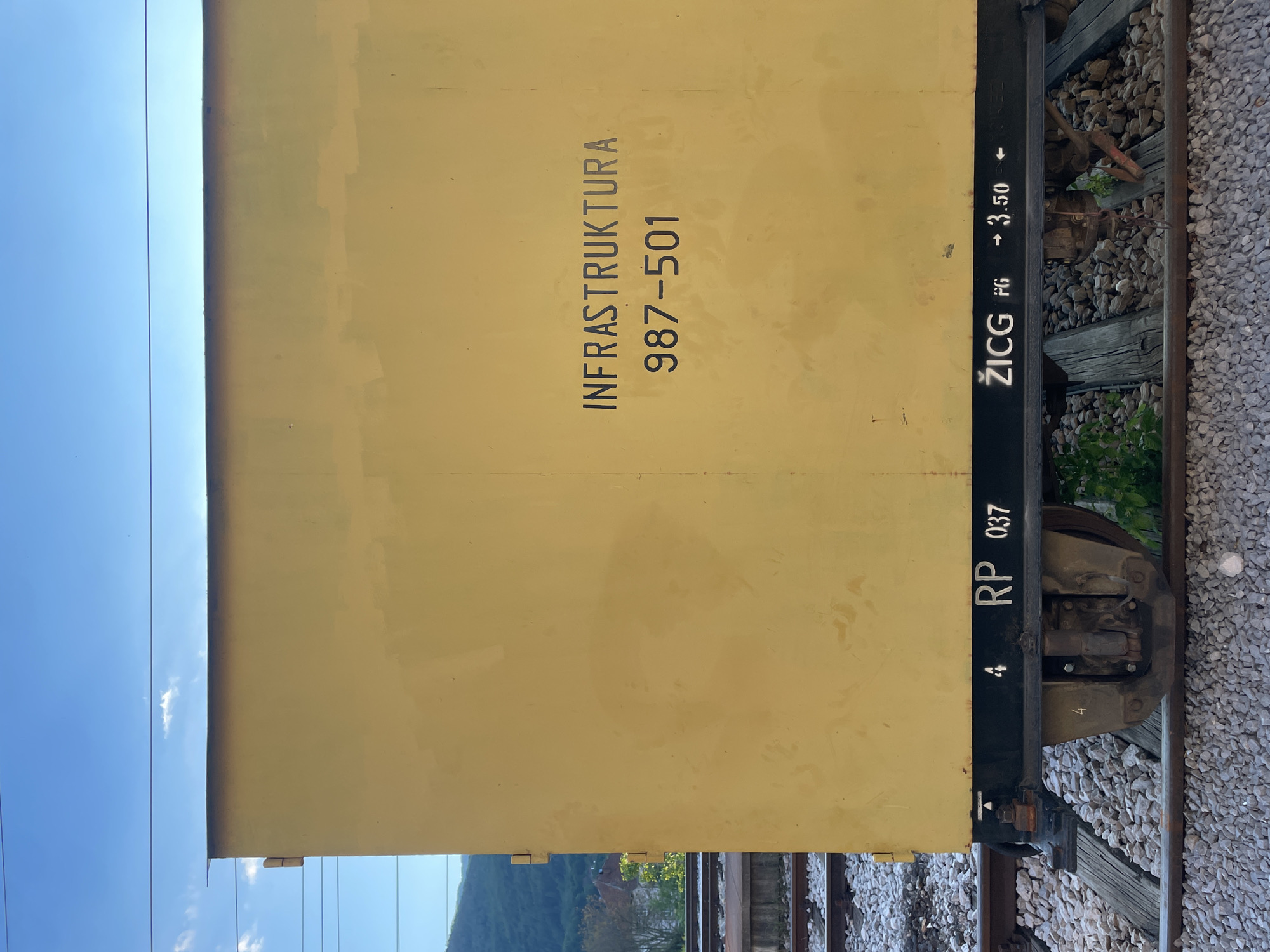 The image depicts a section of a yellow train car, likely photographed in the city of Mojkovac, Montenegro, judging by the word "Infrastruktura" and possibly other elements of the Cyrillic-influenced text. The side of the train car displays "INFRASTRUKTURA" vertically above the number "987-501." Further down, the text "ŽICG" is printed vertically above "FG" with an arrow and "3.50" to the right of "FG," followed by an arrow. On the left side of the image, there is the text "RP 037" vertically. The train car rests on train tracks, which are surrounded by grey gravel. The bottom of the image shows a mountain and a part of the train station. The sky is blue with some thin clouds in the left part of the image.