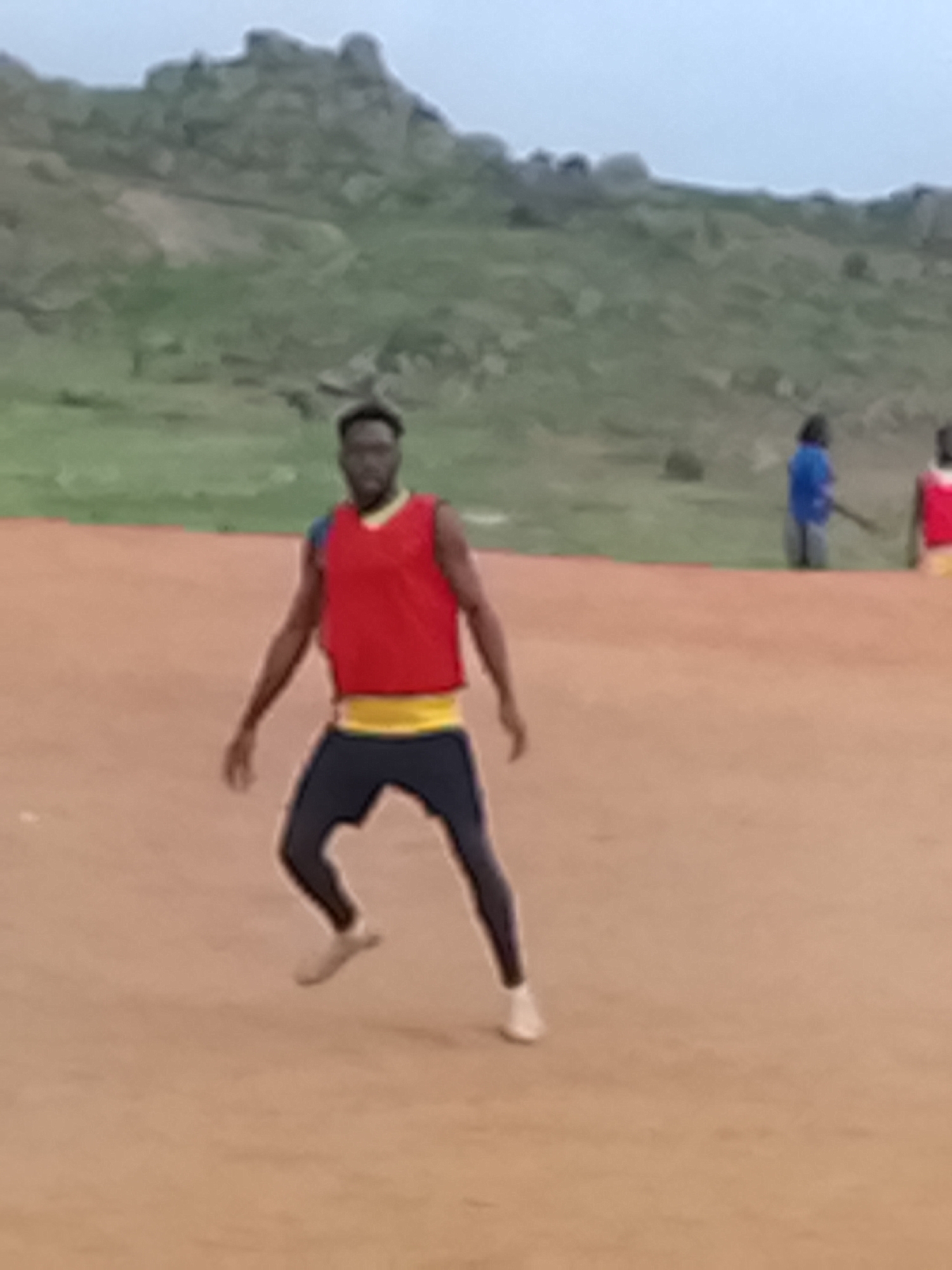 An underexposed, medium-shot image captures a person in athletic attire on a dusty, reddish-brown field. The individual, positioned in the foreground, is wearing a bright red sleeveless vest over a yellow band and dark leggings. Their posture suggests movement, possibly during a sports activity. In the background, a rocky, hilly terrain is visible under a pale sky. Other figures are present further back on the field, one wearing a blue garment. The overall scene depicts an outdoor recreational or sporting event in a natural, arid environment.