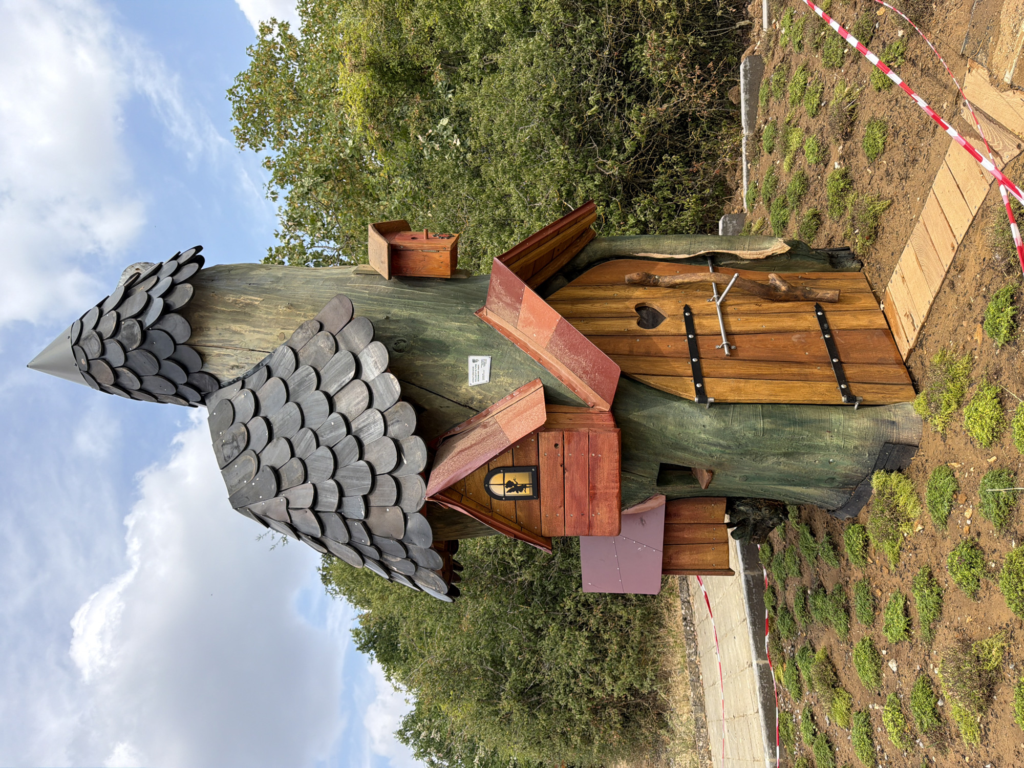 The image shows a whimsical, tree-like structure with multiple small wooden houses and birdhouses attached to its trunk. The structure has a pointed, shingled roof, resembling a witch's hat or a fairy tale dwelling. There is a wooden door with a heart cutout and a latch, as well as several small windows. The structure is situated outdoors, surrounded by greenery and a patch of mossy ground. In the background, there are trees and a cloudy sky. The scene is located in the city of Esch-sur-Alzette, Luxembourg, specifically in the "Stadtwald" (City Forest) area, part of the "Naturpark Öewersauer" (Upper Sûre Nature Park). A path made of wooden planks leads to the structure, and red and white caution tape is visible around the immediate area.