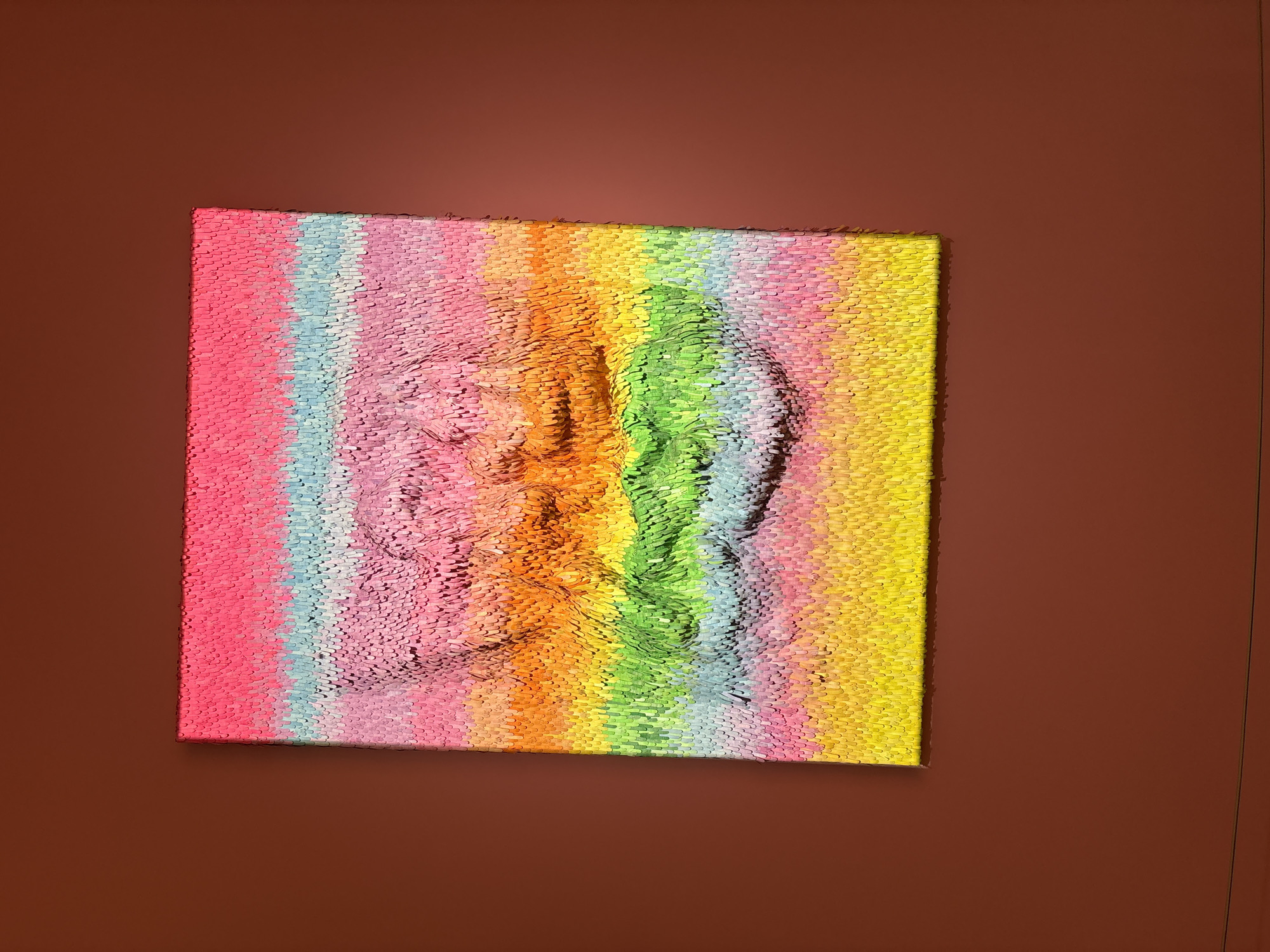 The image shows a square-shaped object hanging on a brown wall. The object appears to be an art piece, potentially a pin impression board or a similar tactile display, with a face pressed into it. The "pins" or elements forming the artwork are colored in a rainbow spectrum, arranged vertically with pink on the left transitioning to yellow on the right, with a green area in the center forming the facial profile. The image displays a three-dimensional effect, creating the impression that the face is emerging from the surface. The brown wall surrounds the artwork, providing a contrasting background. There is no indication of the location being Liangjiang, China, or any other city.