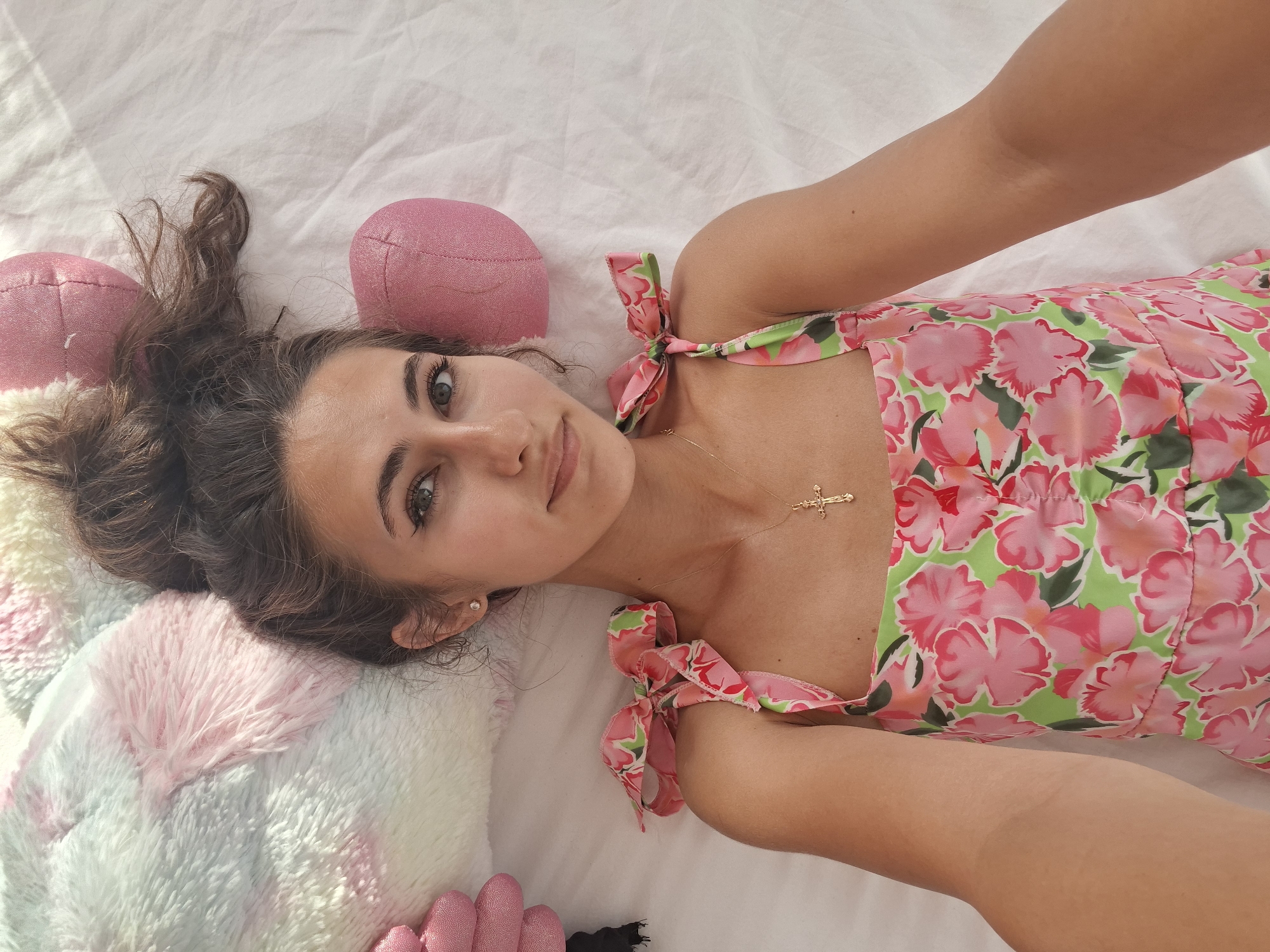 A young woman with dark, messy hair is lying on a white surface, possibly a bed. She is wearing a floral dress with pink and green patterns. A gold cross necklace is visible around her neck. To her left and right are pink, round objects that appear to be pillows or stuffed animals. A large, fuzzy pastel-colored pillow is also on her left. The lighting suggests a bright, indoor setting.
