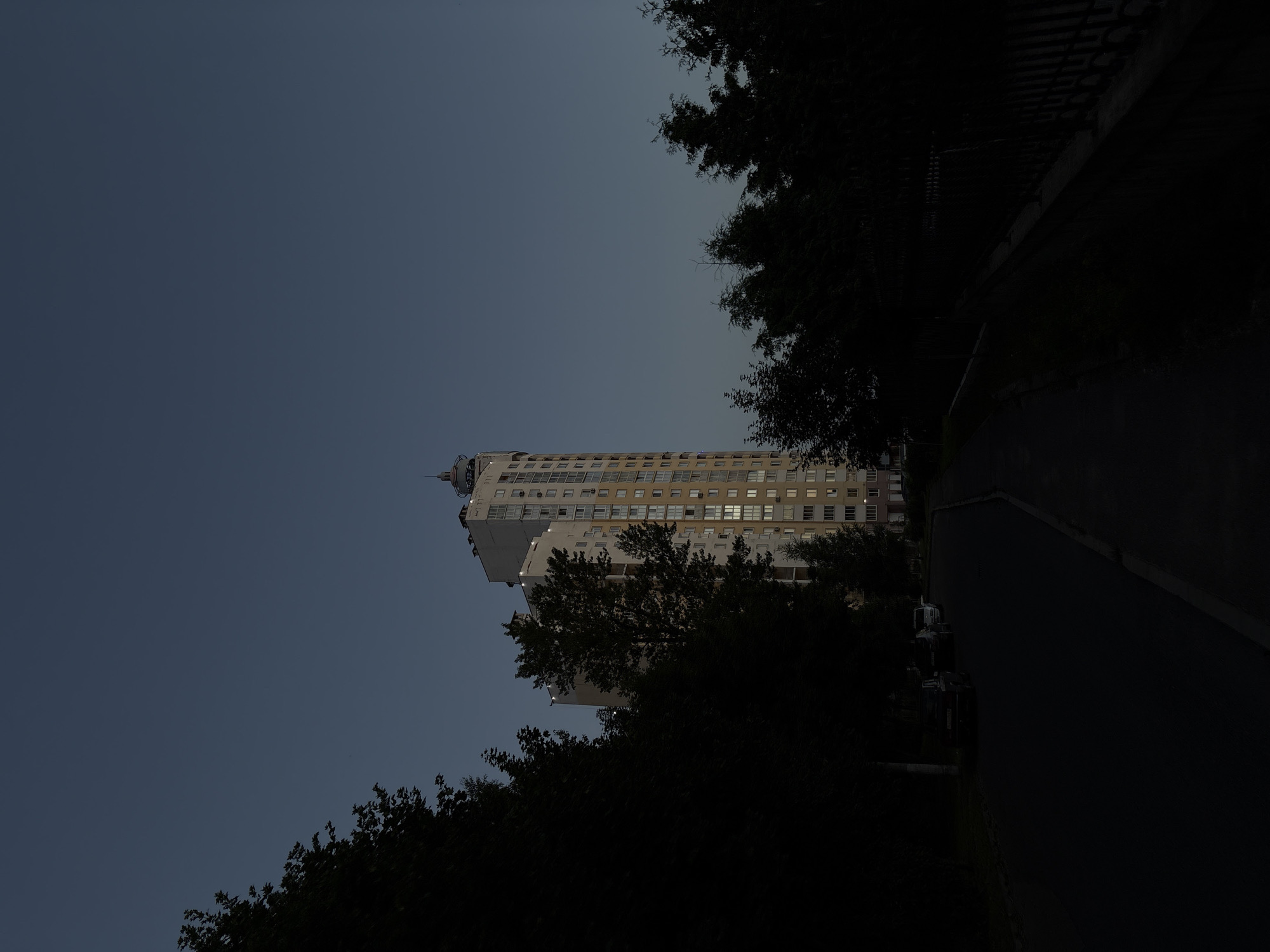 The image depicts a multi-story residential building in Vitsebsk, Belarus, during twilight. The building, with its light-colored facade and numerous windows, stands tall against a darkening blue sky. Some windows are illuminated, suggesting occupants are present. Trees, dense and dark, frame the scene on either side, with their foliage obscuring much of the ground-level surroundings. A paved walkway and a road are visible in the foreground, leading towards the building. Several parked cars are discernible on the road, partially shadowed by the trees. The overall atmosphere is one of dusk settling over an urban residential area.