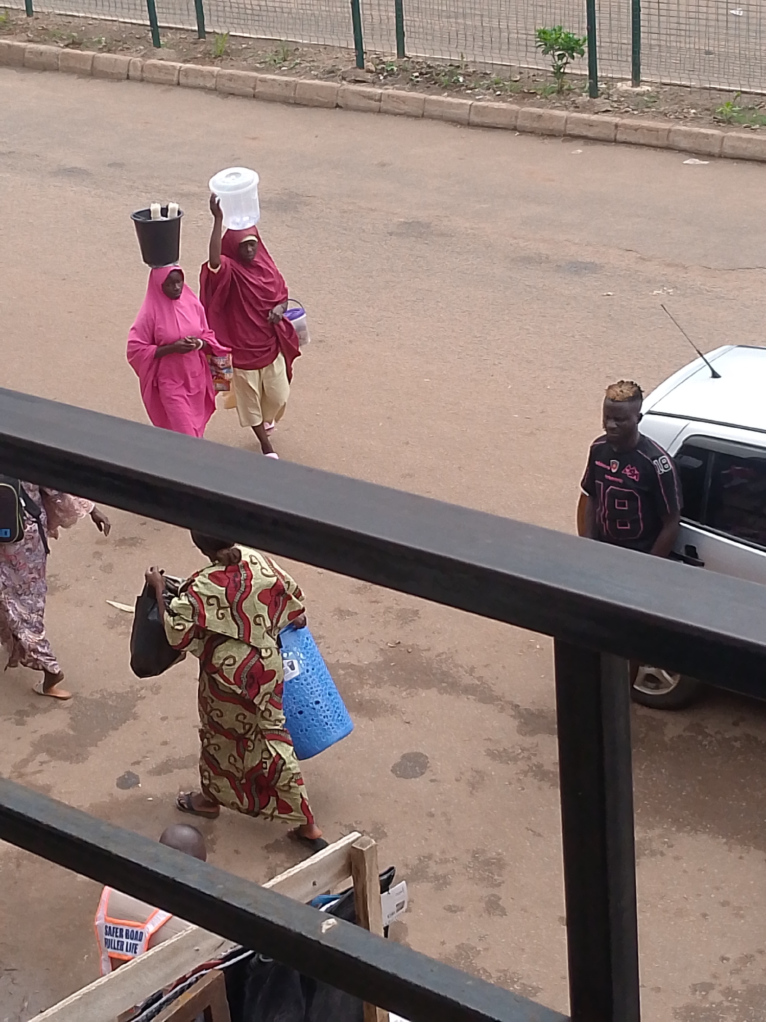 In Bukuru, Nigeria, three individuals are captured in an overhead shot on a paved street bordered by a sidewalk and a metal fence. A woman in a vibrant pink hijab walks with her head balanced by a black bucket containing two white objects. Beside her, another woman, draped in a deep red garment and a lighter beige skirt, carries a large white translucent container on her head and a smaller purple bag in her hand. Further away, a man with styled blond and dark hair wears a dark t-shirt with pink accents and the number "18" prominently displayed, standing near a silver car. To the left, a fourth person, partially obscured by a dark railing in the foreground, wears a patterned dress and carries a black bag and a blue basket. Another individual, wearing a high-visibility vest that reads "SAFER ROAD FULL LIFE," is partially visible at the bottom. The scene is outdoors, with the ground showing patches of dampness and scattered debris.