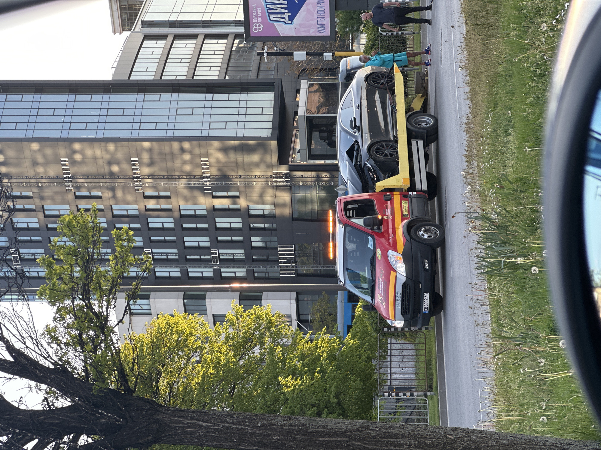 A red and yellow tow truck, license plate CB5952AO, is stopped on the side of a street in Sofia, Bulgaria. On the tow truck is a grey passenger car with damage to the front end. Two individuals are standing near the tow truck. A tall building with a billboard is in the background. Grass and dandelions are along the side of the road. A tree trunk is visible in the lower left of the image, and a car mirror is visible on the right.