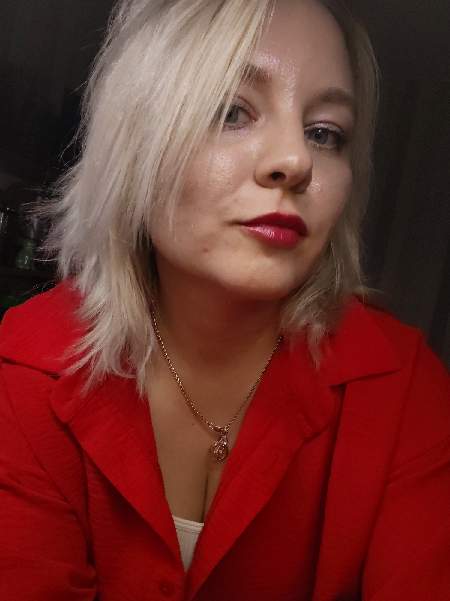 A fair-skinned woman with blonde, shoulder-length hair is facing the camera. She has blue eyes and red lipstick. She is wearing a red collared shirt and a gold necklace with a pendant. The background is dark and indistinct, with some blurred green objects visible in the upper left. The location is not identifiable as Izhevsk, Russia.
