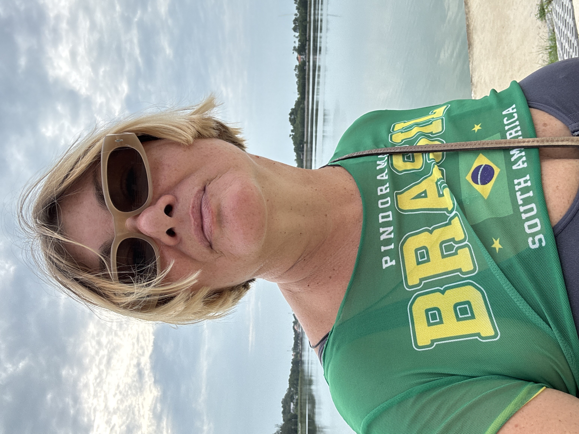 A selfie shows a woman with short blonde hair and tan-framed sunglasses in front of a body of water. She is wearing a green shirt with "BRASIL" written in yellow, along with the words "PINDORAMA" and "SOUTH AMERICA," along with the flag of Brazil. The background appears to be a cloudy sky and a lake or river with vegetation on the opposite shore. The location is likely บ้านบางรักษ์, Thailand. The woman is facing the camera and the shirt is the most prominent feature of the photograph.