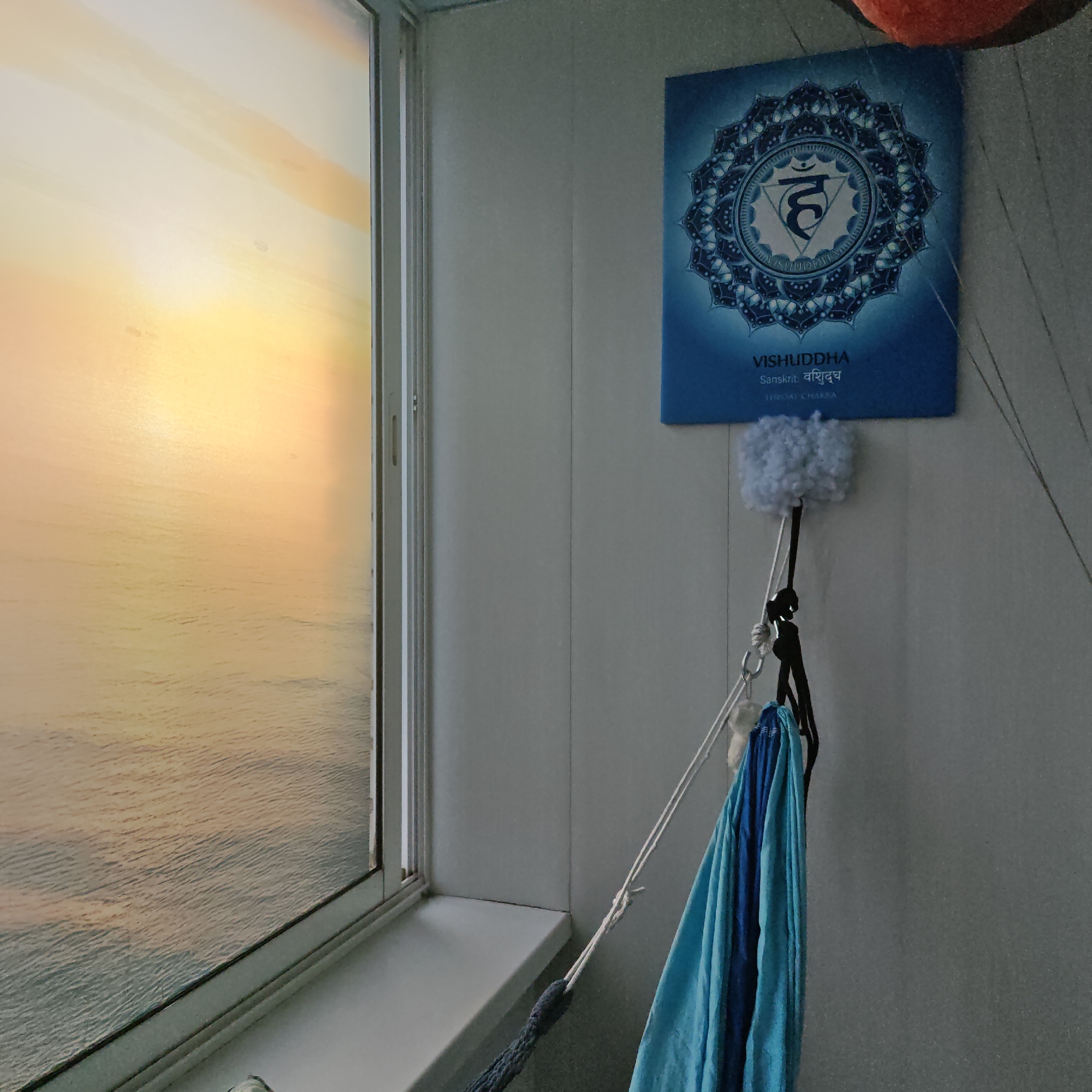 The image depicts an interior scene in Kladovitsy, Russia. To the left, a window is visible, revealing a view of water reflecting sunlight, possibly an ocean or a large lake at sunset. The window frame is white. Beside the window, a white wall is decorated with a blue poster displaying a mandala-like image with the text "VISHUDDHA" and the word "Chakra" beneath. Hanging from the wall below the poster is a device with a fuzzy white component, ropes, and a metal clip, supporting a draped blue fabric. The overall lighting suggests it is either early morning or late evening.
