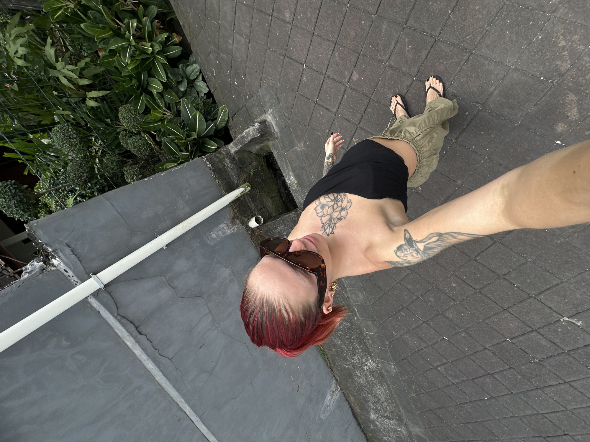 A woman with bright red hair is taking a selfie outdoors, likely in North Kuta, Indonesia. She is wearing large, tortoiseshell-patterned sunglasses, a black tube top, and loose-fitting olive green pants. Her left arm, visible in the foreground, features a large tattoo of a bird and floral designs. Her right arm, also tattooed, is visible at her side. She has visible tattoos on her chest as well. The ground is paved with dark gray square tiles. To her left, a rough, gray concrete wall runs along the frame, with a white pipe attached to it. To her right, a lush green garden is visible behind a wire fence, with various plants and trees. The scene appears to be during the daytime, with overcast or diffused lighting.