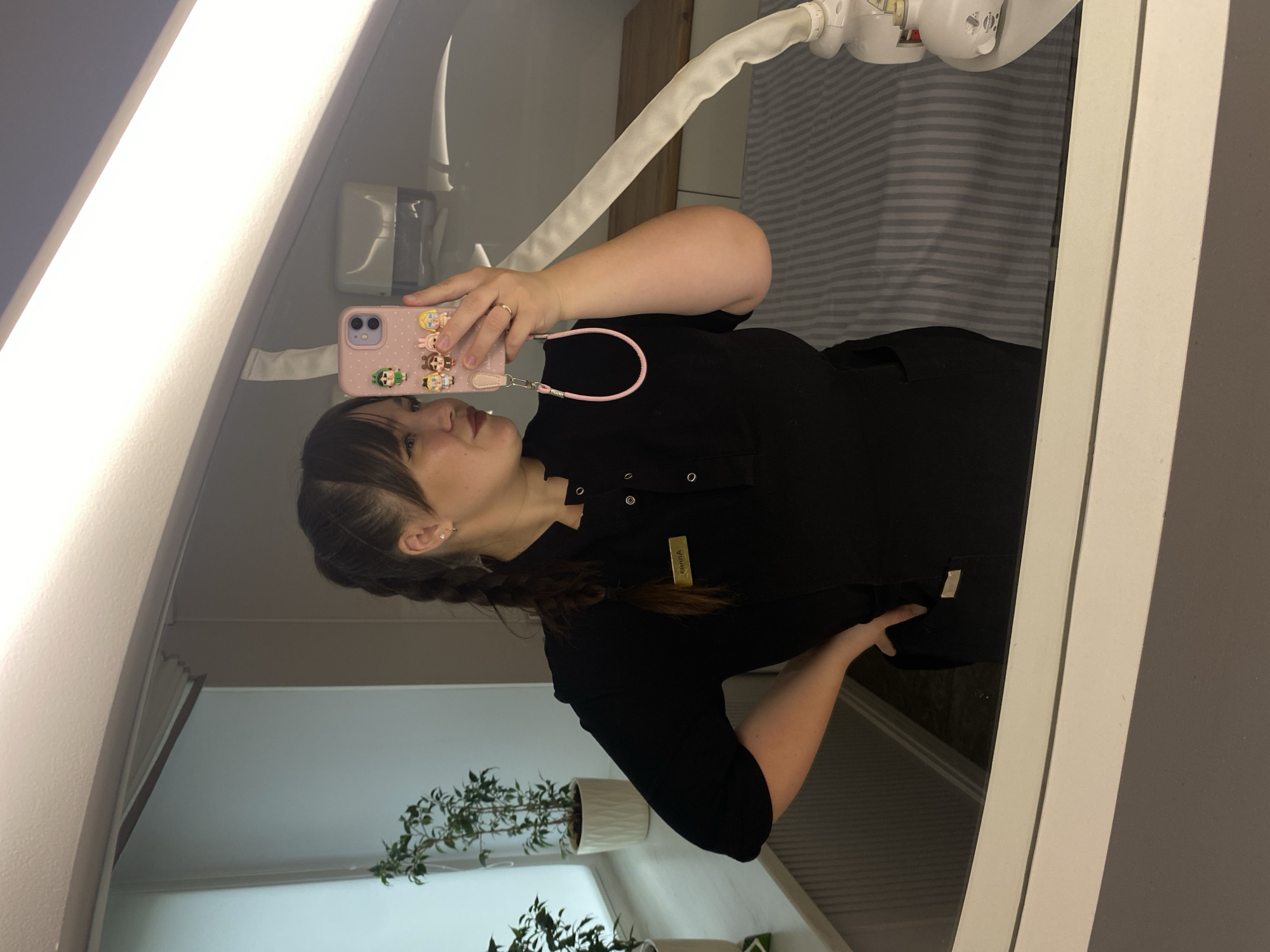 A woman is taking a mirror selfie in what appears to be a medical or spa setting. She is wearing a black scrub top with a name tag that reads "SARA". Her hair is in a braid and she is holding a pink phone case adorned with cartoon characters. In the reflection, we can see a sink, a striped curtain, and a light fixture. There are also two potted plants visible on a shelf in the lower part of the reflection. The scene appears to be indoors, likely in a professional environment. The lighting suggests it could be daytime, but it's hard to tell definitively. The woman's expression is neutral, and the overall mood is casual and everyday.