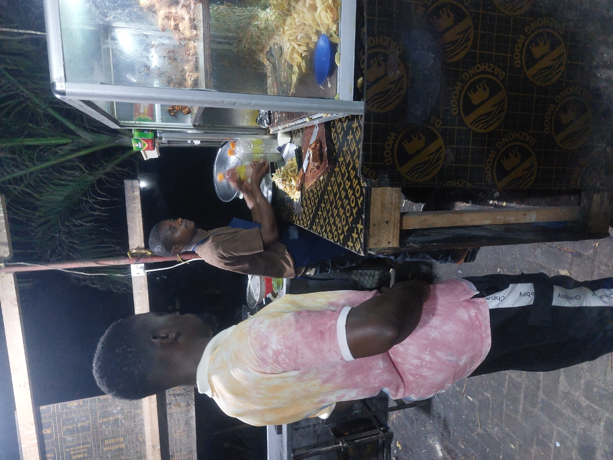 Here is a description of the image:
**Setting:** The image appears to be taken at a street food stall in Dar es-Salaam, Tanzania, at night.
**People:** There are two African men in the image. One man is standing behind the food stall, preparing food. He is wearing a brown shirt and a blue apron. The other man is standing in front of the food stall. He is wearing a pink and yellow t-shirt, and black pants with white branding.
**Objects:** The main object in the image is the food stall. The stall has a glass display case containing french fries and fried chicken. There are also bottles of condiments, food preparation items, and other food-related objects on the counter. The side of the stall is made of dark plywood with a repeated "DAZHONG WOOD" logo. There are some palm fronds visible in the background.
**Actions/Interactions:** The man behind the counter is actively preparing and serving food, he's plating the food into the containers. The other man in front might be waiting for an order.