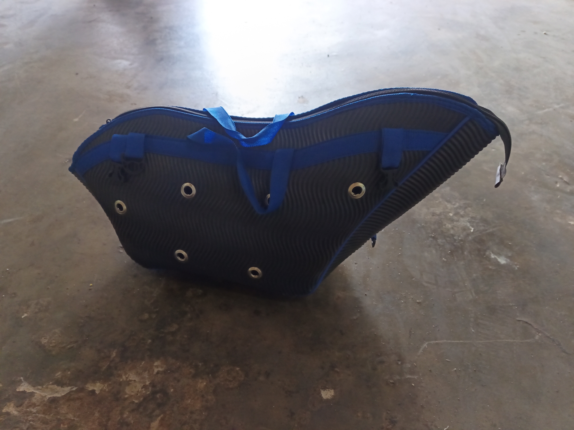 A dark blue, contoured bag with bright blue straps and trim rests on a mottled grey concrete surface. The bag has a zippered opening along its top edge and features several circular metal grommets or eyelets on its body. The concrete floor exhibits wear and tear, with patches of lighter and darker discoloration, and scattered small debris. The overall lighting suggests an outdoor or well-lit indoor environment. The image is captured from a slightly elevated, oblique angle, looking down at the bag. There are no people or animals visible in the scene. The location of Terbanggi Marga, Indonesia, is not visually represented in the image itself.