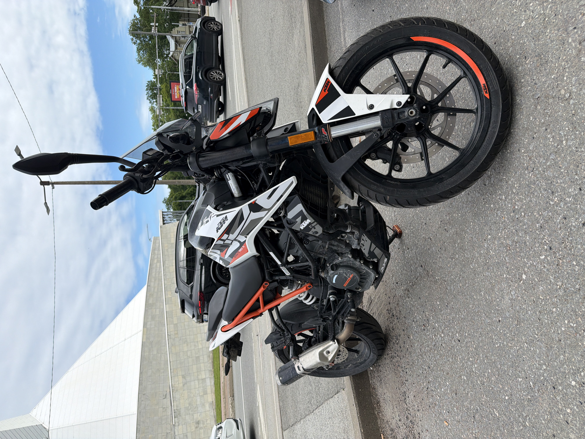 A white and black KTM Duke motorcycle is parked on a paved street in Tallinn, Estonia. The motorcycle has orange accents on its frame and wheels. In the background, there is a modern white building with a geometric design, and several cars are driving on the street. The sky is blue with some clouds. The scene appears to be during the daytime.