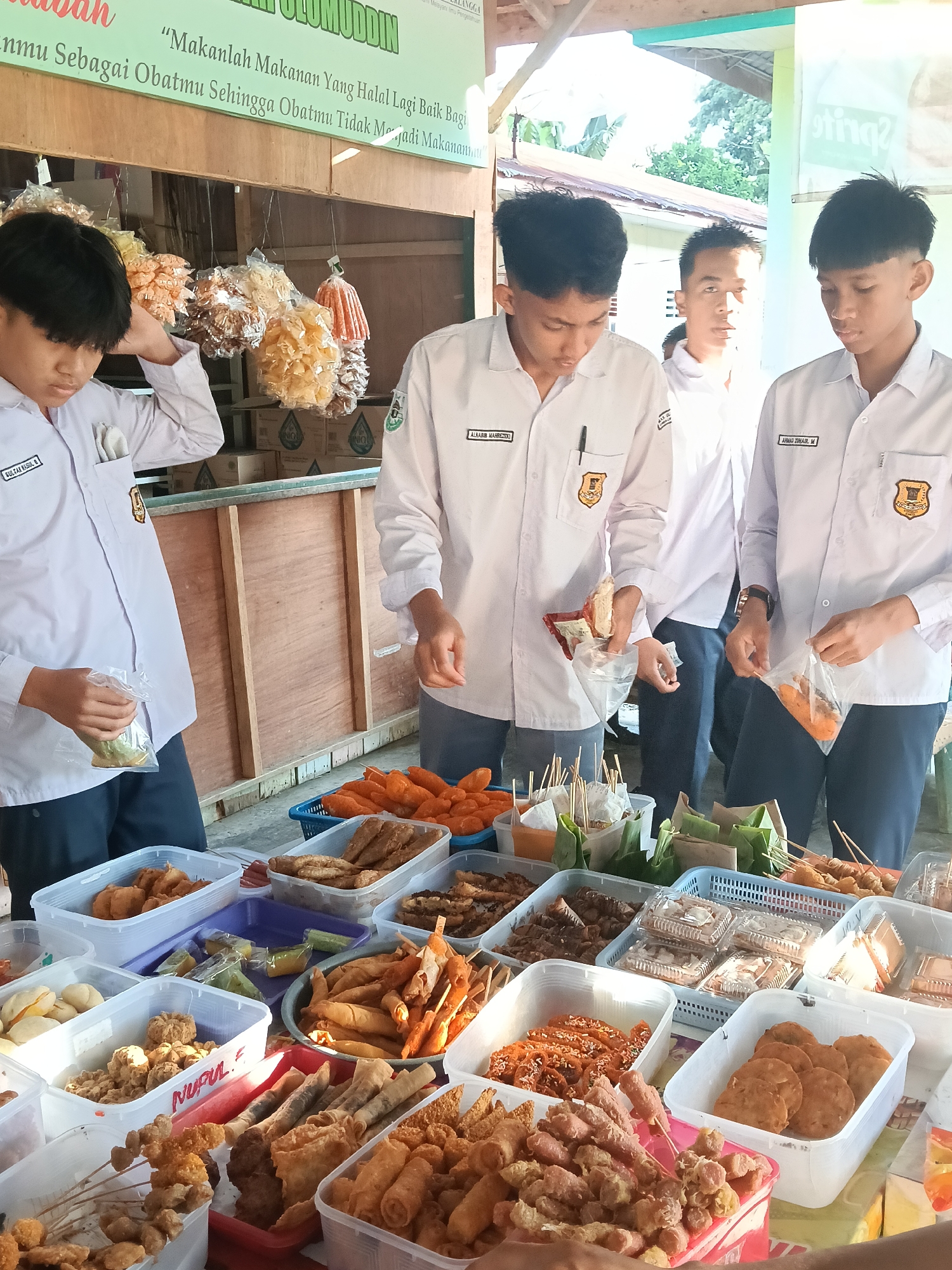 Captured in Lhokseumawe, Indonesia, the image showcases four young students in white uniform shirts, presumably on a school break, standing at a food stall. The stall is laden with various fried snacks and skewers in plastic containers. The students are examining the food and some have already selected items, holding them in plastic bags. One student is touching his hair while holding a bag. A banner with Indonesian text is visible above the stall.