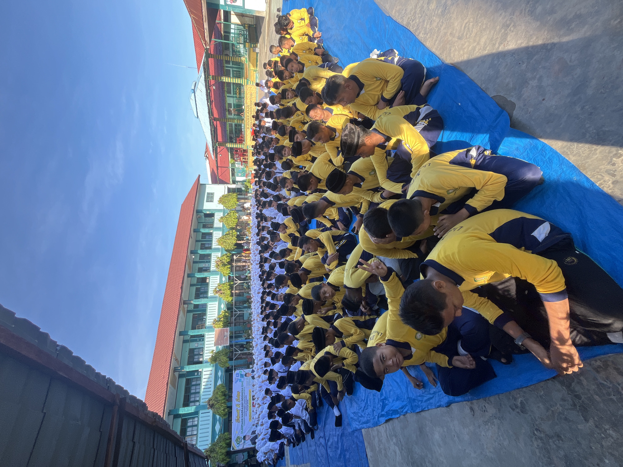 In Lhokseumawe, Indonesia, a group of male students, predominantly wearing yellow long-sleeved shirts and navy blue pants, are seated on blue tarpaulins outdoors. The students, of various ages, are arranged in rows, facing away from the camera, with some in the foreground looking towards the viewer. One student in the foreground is making a peace sign with his fingers. Behind them, more students in similar attire are visible, forming a large gathering. In the background, a building with a red roof and light blue facade stands, suggesting a school or educational institution. A banner with text is displayed in front of the building. The scene is set under a clear blue sky with bright sunlight casting shadows.