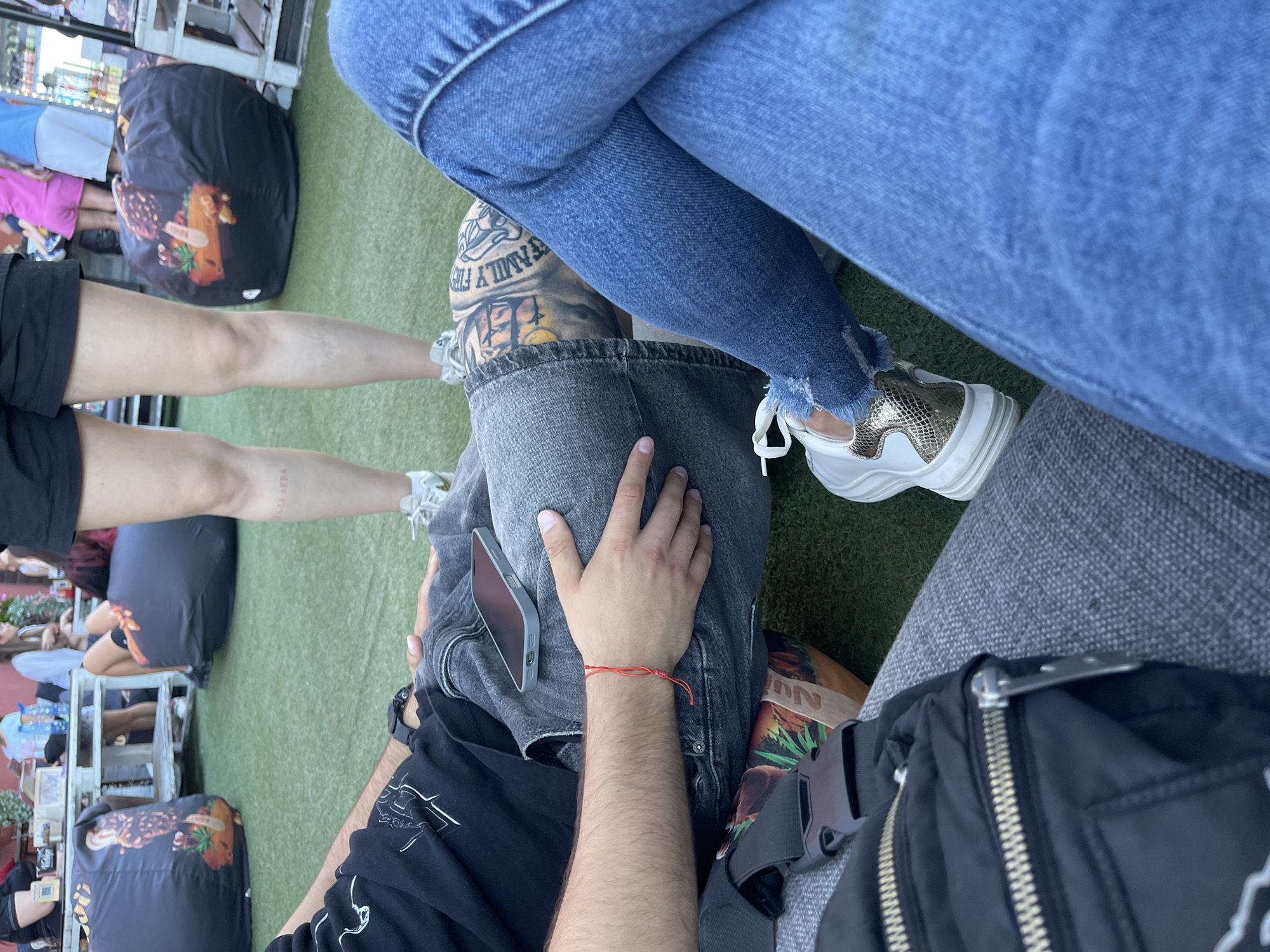 In an outdoor setting with green artificial grass, two individuals are seated. The person on the left, wearing a black hoodie and gray jeans, has a red string bracelet on their left wrist and a smartphone resting on their lap. Their arm is resting on their leg. The person on the right is wearing blue jeans and white sneakers with gold accents. Visible tattoos are present on the left leg of the person on the left. In the background, several large black beanbag chairs with graphic prints and a white table are scattered. Other individuals are present in the background, some standing and some seated, with various beverages on shelves behind them. The lighting suggests it is daytime.