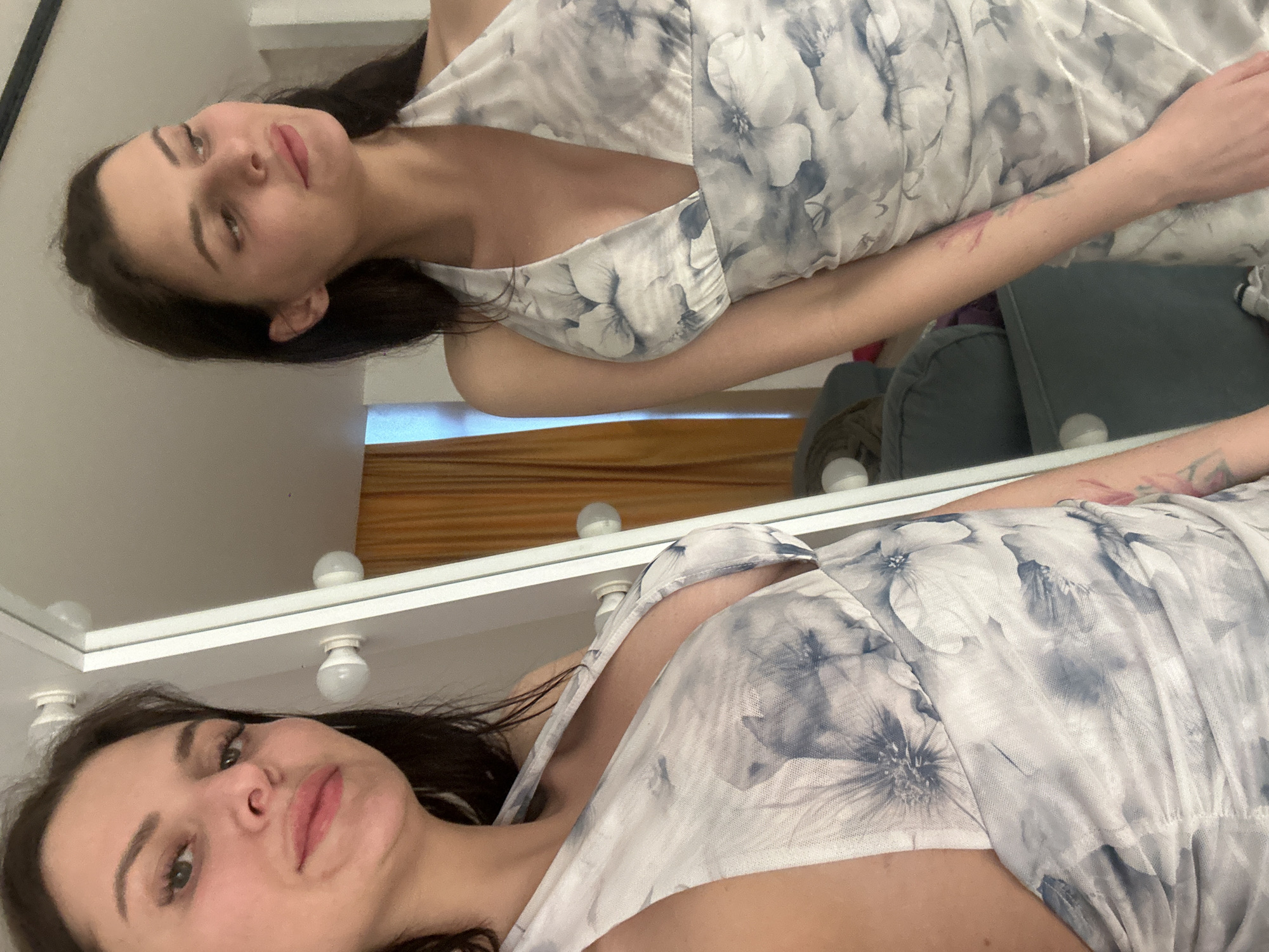 A woman is captured in a mirror selfie, her reflection showing her to the right and herself to the left. She is wearing a white halter dress with a floral pattern in shades of blue and gray. The dress has a deep V-neckline. The woman has dark hair and is looking slightly away from the camera with a soft smile. Her left arm is visible, and it appears to have a tattoo. The background shows a room with a mirror frame that has lights around it, suggesting a vanity or dressing area. There are curtains in a warm yellow hue behind the woman, and a gray sofa is visible in the reflection. The overall setting is indoors, likely a domestic environment.