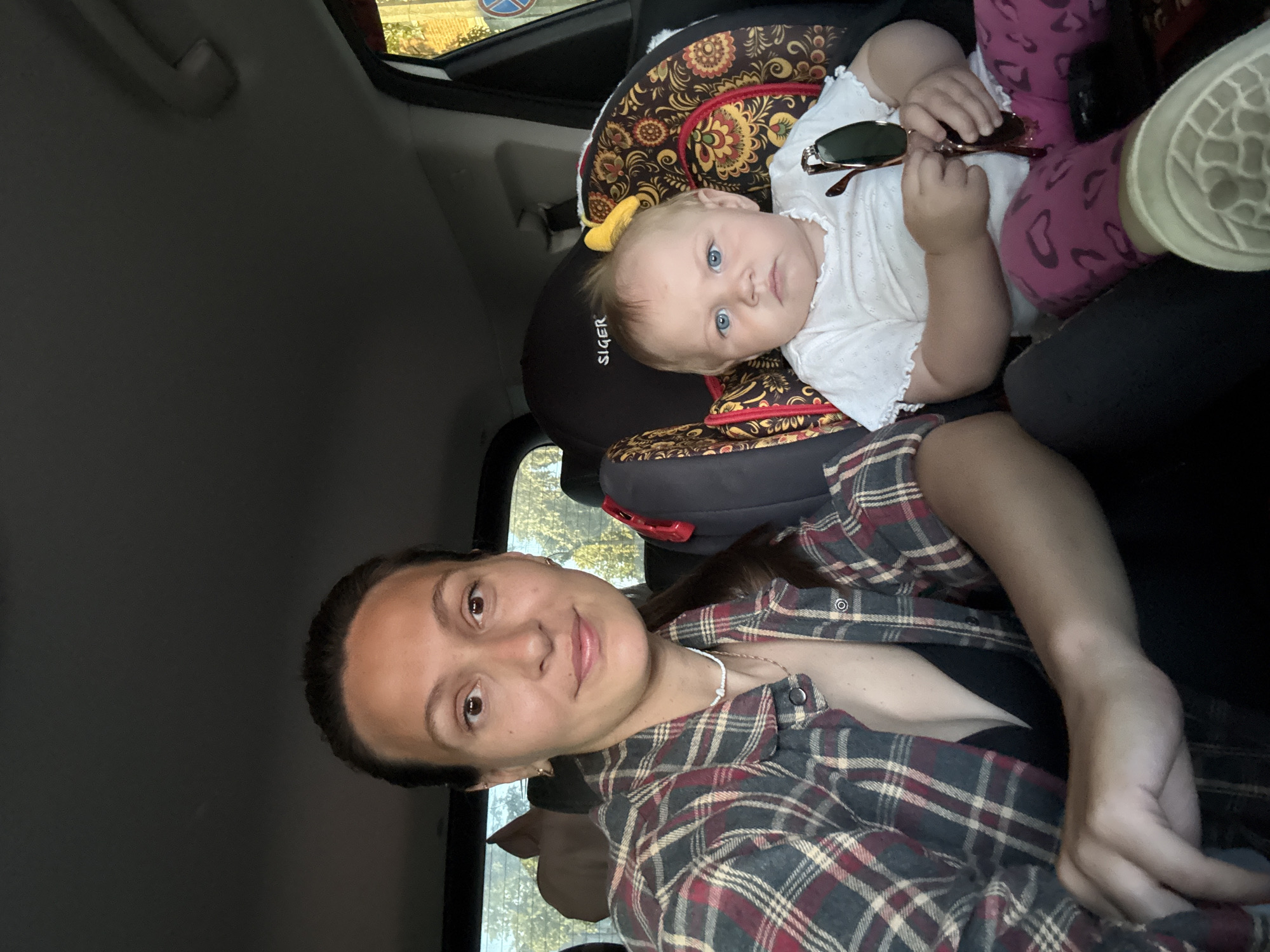 A woman and a baby are in the back seat of a car. The woman is on the left, wearing a plaid shirt and a black bra. The baby is in a car seat on the right, wearing a white top and pink leggings. The baby is holding a pair of sunglasses. The car appears to be in an outdoor setting, with trees visible through the windows. The scene is likely in Chaikovsky, Russia, given the provided location.