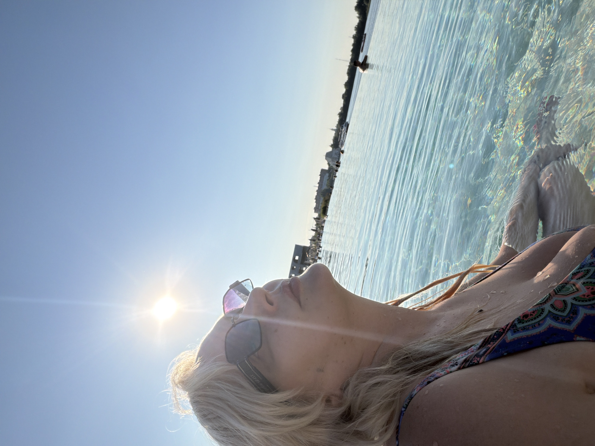 A woman with blonde hair and sunglasses is in the water, looking up at the sun. She is wearing a patterned swimsuit. The sun is bright and shining in the clear blue sky. In the background, there is a body of water with people swimming and boats on the horizon. There are buildings and trees along the shoreline. The water is clear and blue, with sunlight reflecting off the surface.