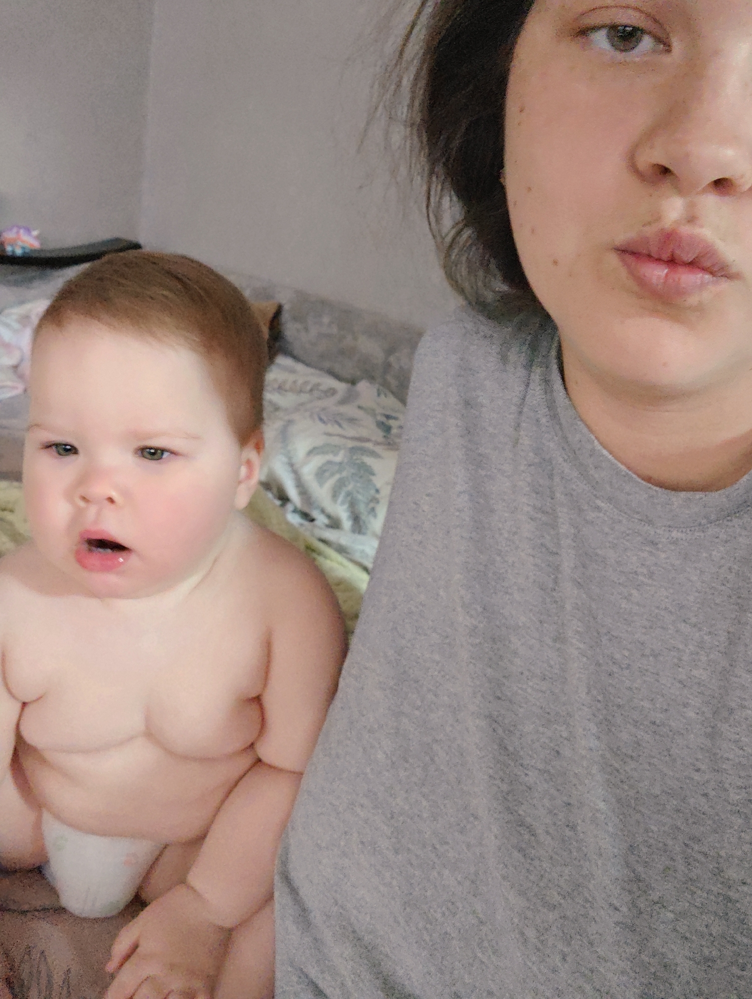 This close-up portrait, captured in Chaikovsky, Russia, features an adult and a baby in an indoor setting. On the right side of the frame, an adult, likely female, is partially visible from the nose down to the chest. She has dark hair, light-colored eyes, and freckles on her cheek. Her lips are slightly puckered, and she is wearing a plain grey t-shirt.
To the left, a baby is depicted from the head to mid-thigh. The baby has light brown hair, light eyes (possibly green or hazel), and a somewhat surprised or curious expression with its mouth slightly open. Its rosy cheeks and visible body rolls suggest a plump infant. The baby is bare-chested and wears a white diaper.
The background shows a plain grey wall and a bed covered with a light-colored blanket or bedspread featuring subtle light blue/grey leaf or floral patterns. A dark, narrow object, possibly a shelf, is partially visible in the upper left corner, holding a small, colorful object that appears to be a unicorn toy. The image captures a casual, intimate moment, likely a selfie, with no specific activity beyond posing for the photo. The time of day is not evident from the indoor lighting, and there is no visible text.