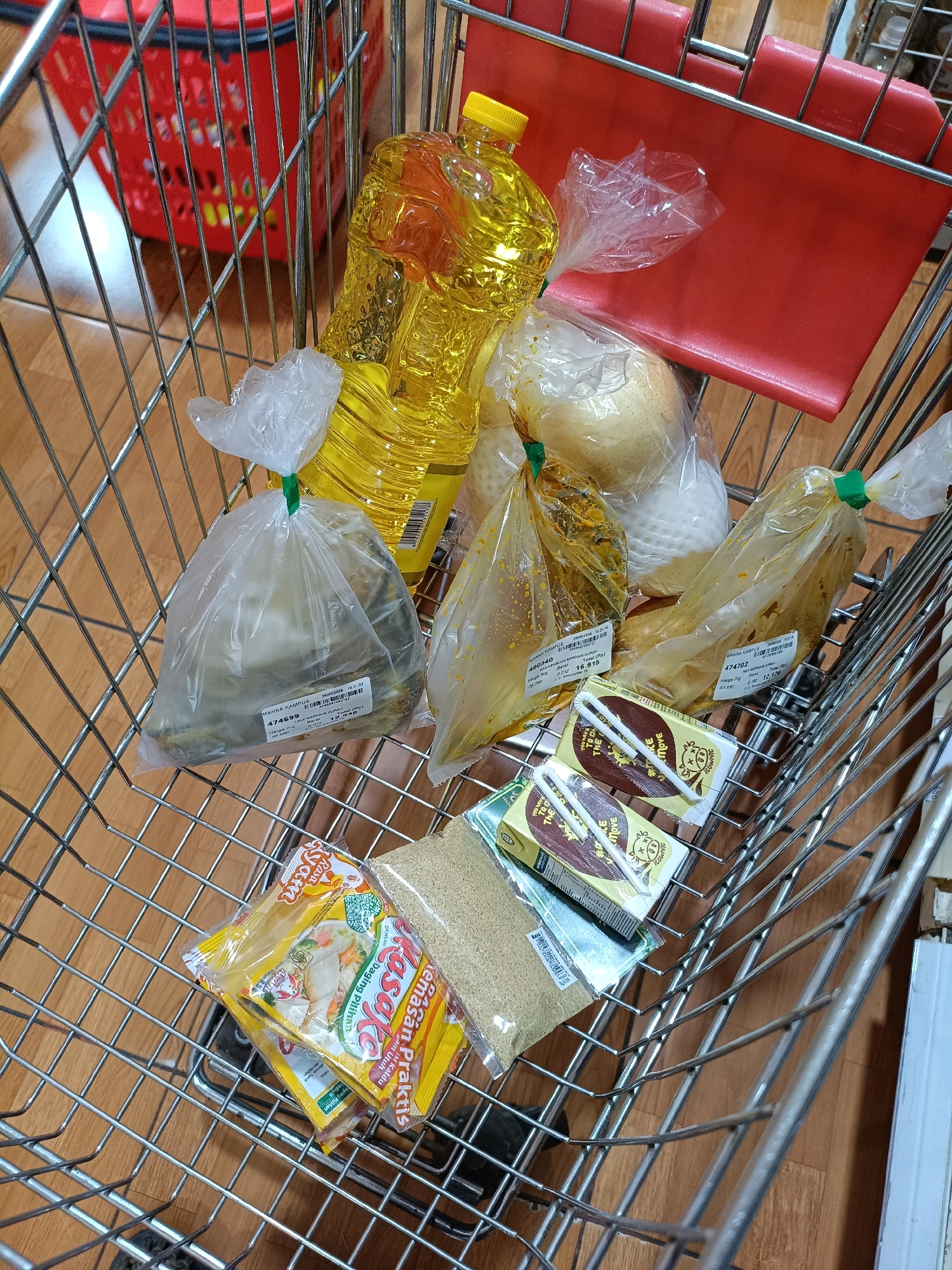In a shopping cart at a supermarket in Yogyakarta, Indonesia, are various food items. A large bottle of cooking oil with a yellow cap is prominent. Several plastic bags contain food items, including one with what appears to be fish and others with pre-cooked dishes in yellow curry sauce. Two individually packaged pears are also visible. At the bottom of the cart, there are boxes of instant noodles and a packet of what looks like ground spices. The shopping cart is made of metal and is on a tiled floor. In the background, a red plastic basket is attached to the cart.