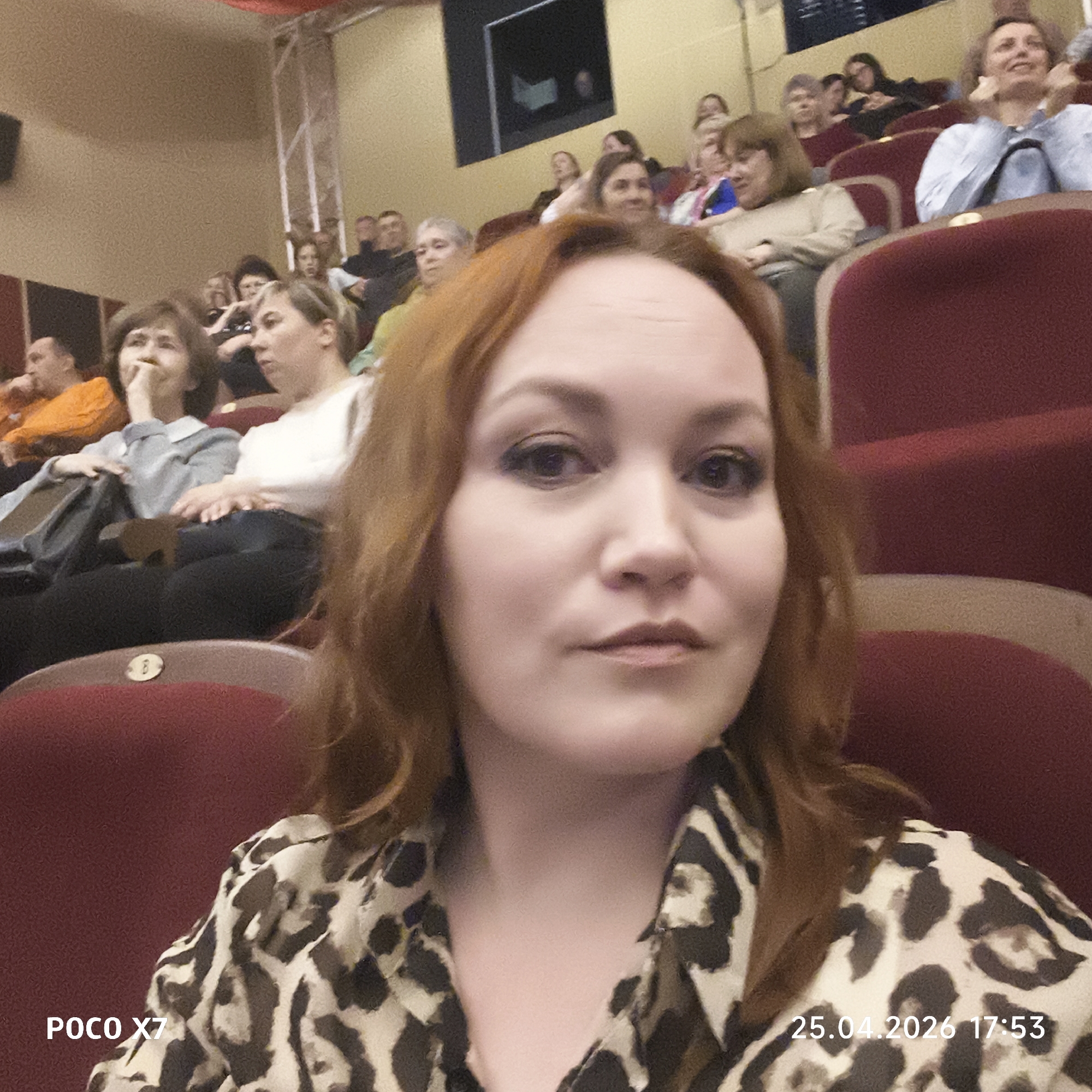 The image depicts a selfie taken by a woman with reddish-brown hair and a leopard-print shirt in what appears to be a theater or auditorium. She is seated in a red cushioned seat. Other people are seated in rows behind her. The timestamp indicates the photo was taken on April 25, 2026, at 17:53. Based on the context, this may have been taken in the city of Chaikovsky, Russia.