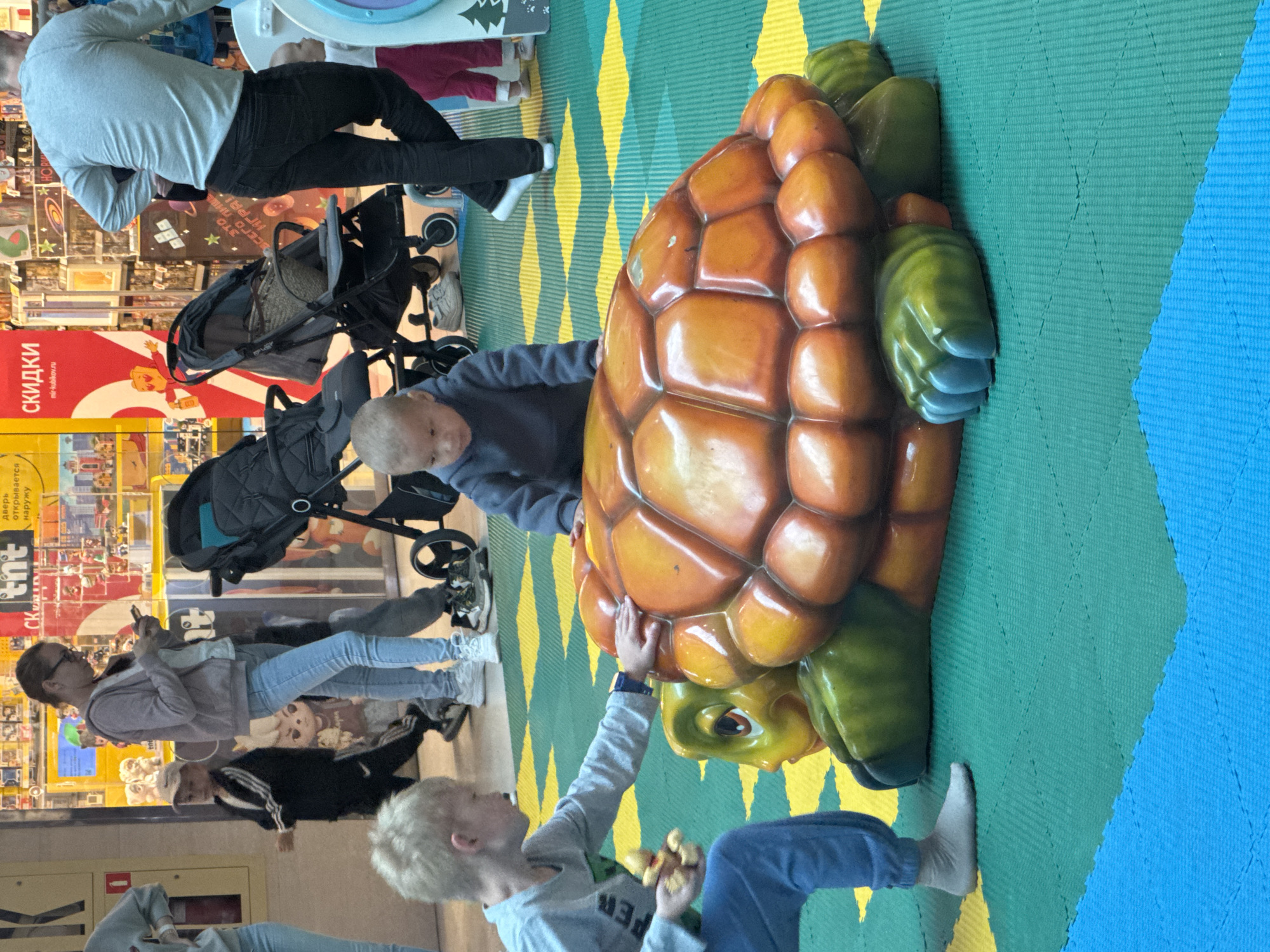 This image captures an indoor scene, likely a children's play area or shopping mall, with a large, colorful turtle sculpture as the central focus. A young boy with blond hair, wearing a gray long-sleeved shirt and blue pants, is interacting with the sculpture, holding a small yellow toy. To his right, another young child, dressed in a blue hooded sweatshirt, is seated near the turtle. In the background, several adults and children are visible, along with shopping displays, strollers, and what appears to be a retail environment. The floor is covered with a mat composed of interlocking blue, green, and yellow sections. The overall impression is one of a casual, public space designed for families and children. There is no specific indication of the city of Ufa, Russia, within the image itself.