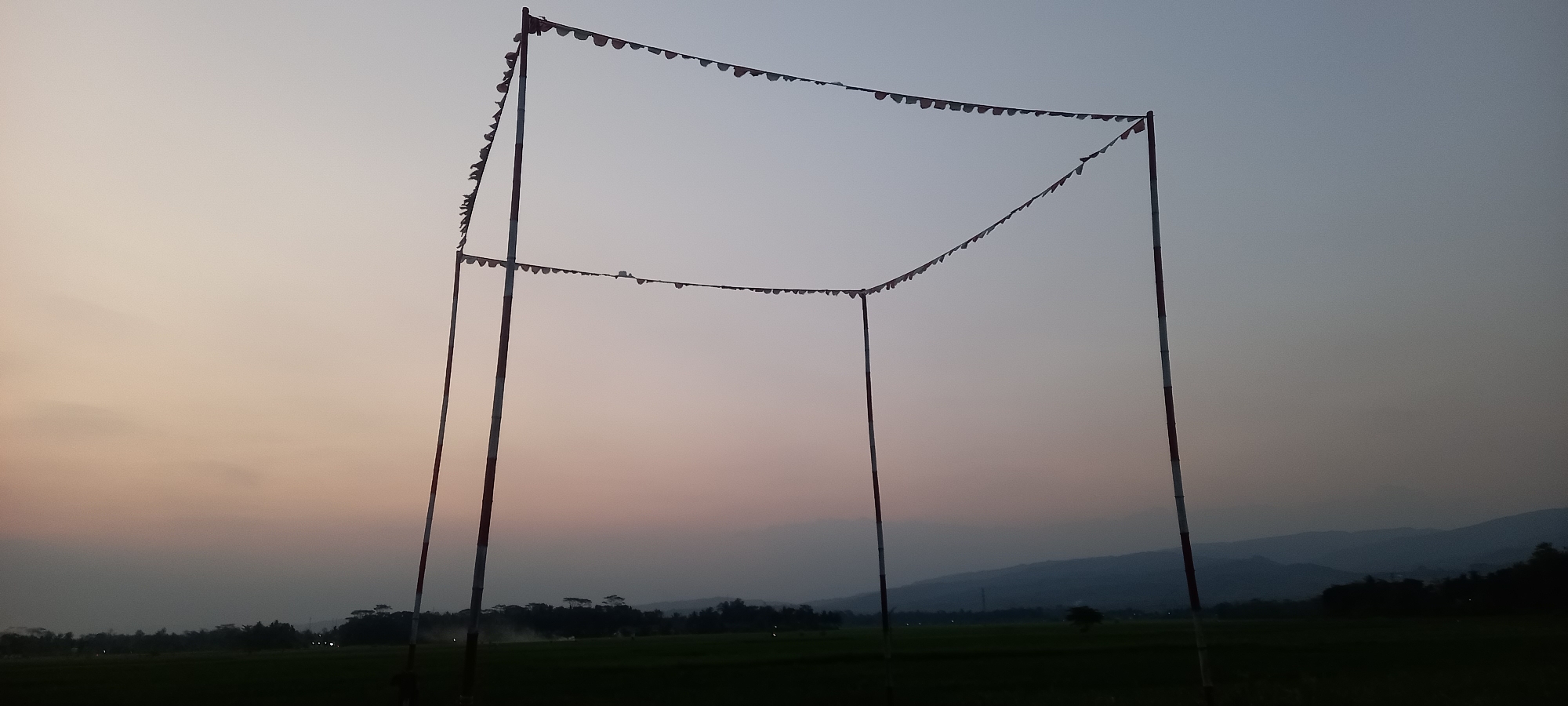 In Sidamulya, Indonesia, at dusk, a rectangular frame structure made of four poles adorned with small red and white flags stands in a dark, open field. The flags are strung along the top and middle horizontal supports of the frame. The sky exhibits a soft gradient of colors, transitioning from pale yellow near the horizon to a muted blue overhead, indicating the fading light of sunset. In the background, silhouetted trees and rolling hills are visible against the dusky sky. The overall atmosphere is serene, with no people or animals present.