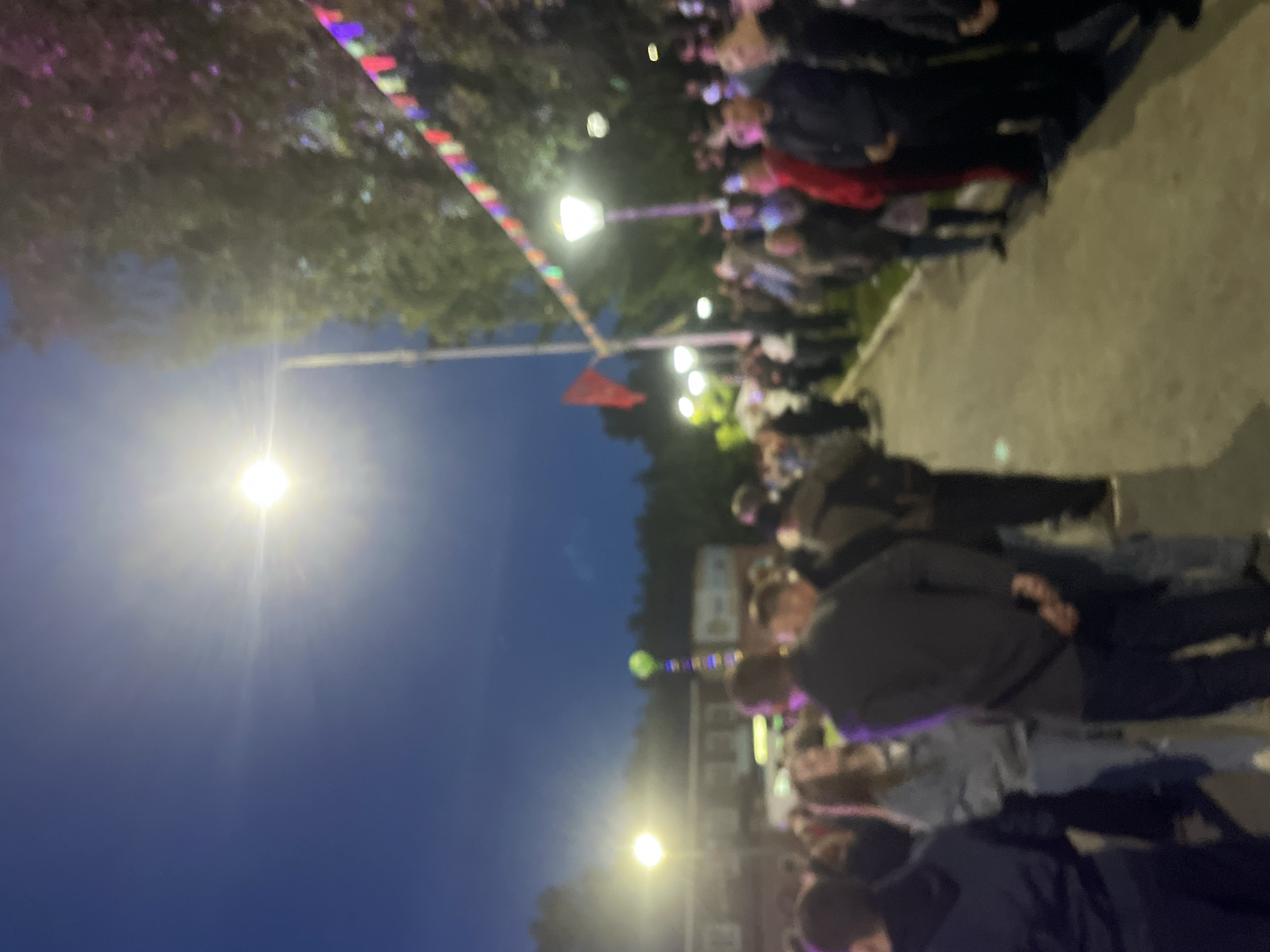 The image depicts an outdoor event in what appears to be the city of Красные Баки, Russia. A crowd of people is gathered, illuminated by streetlights and festive colorful lights strung above. Some people are wearing casual clothing, and others are wearing more formal attire. A red flag can be seen hanging. The environment suggests a public space, potentially a square or street during an event or celebration.