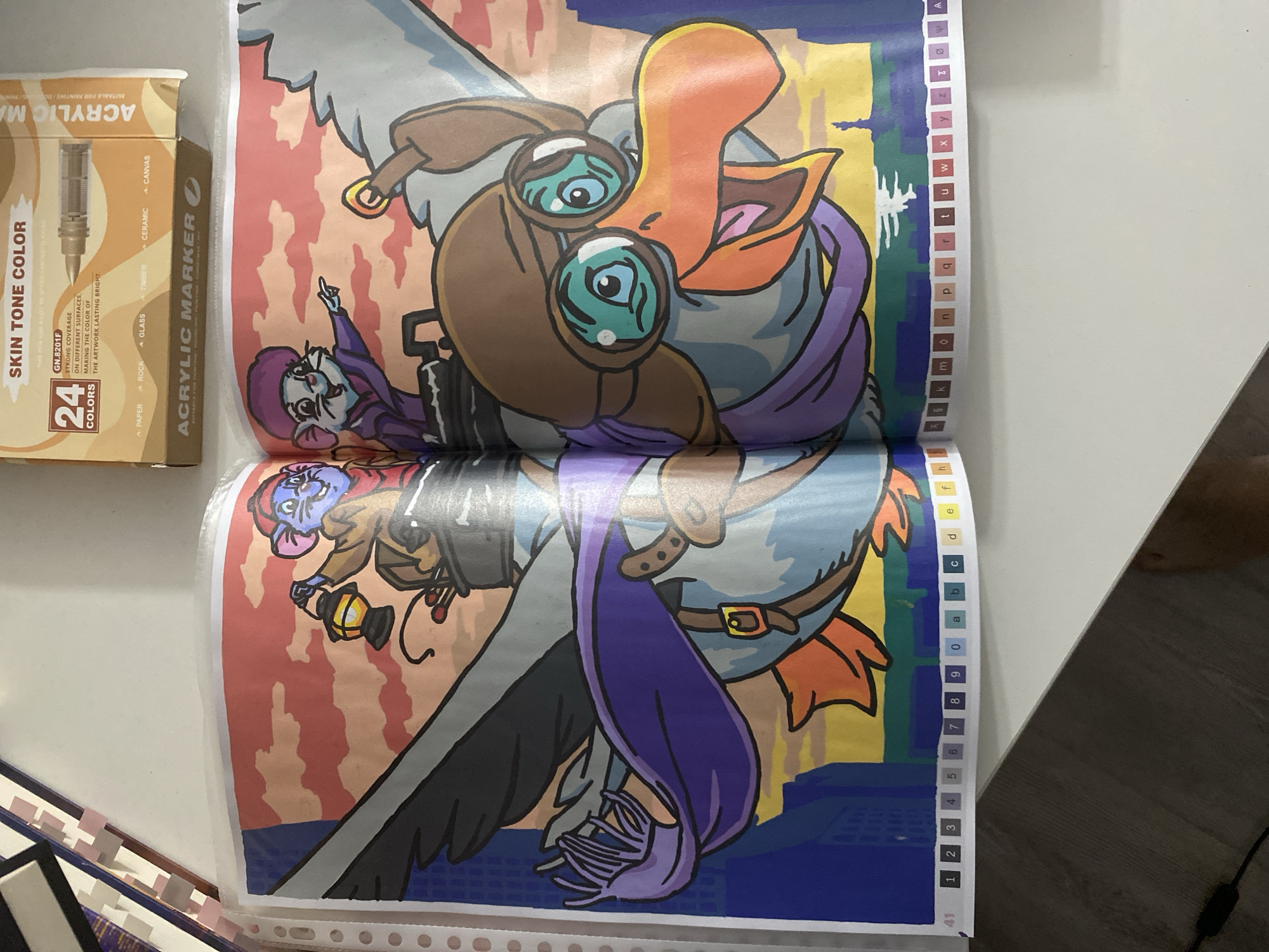 The image depicts a coloring book page with a cartoon illustration of two mice and a duck piloting an aircraft. The mice are dressed in aviator attire, with one holding a lantern and the other pointing forward. The duck, also in pilot gear, has large goggles and an open beak. The background shows a stylized cityscape with a sunset sky. Along the bottom edge of the page, there is a numbered and lettered key, typical of a paint-by-numbers or coloring book. The image is taken in an indoor setting, with a white table surface visible. There are no discernible elements in this image that specifically identify its location within the city of Łódź, Poland.