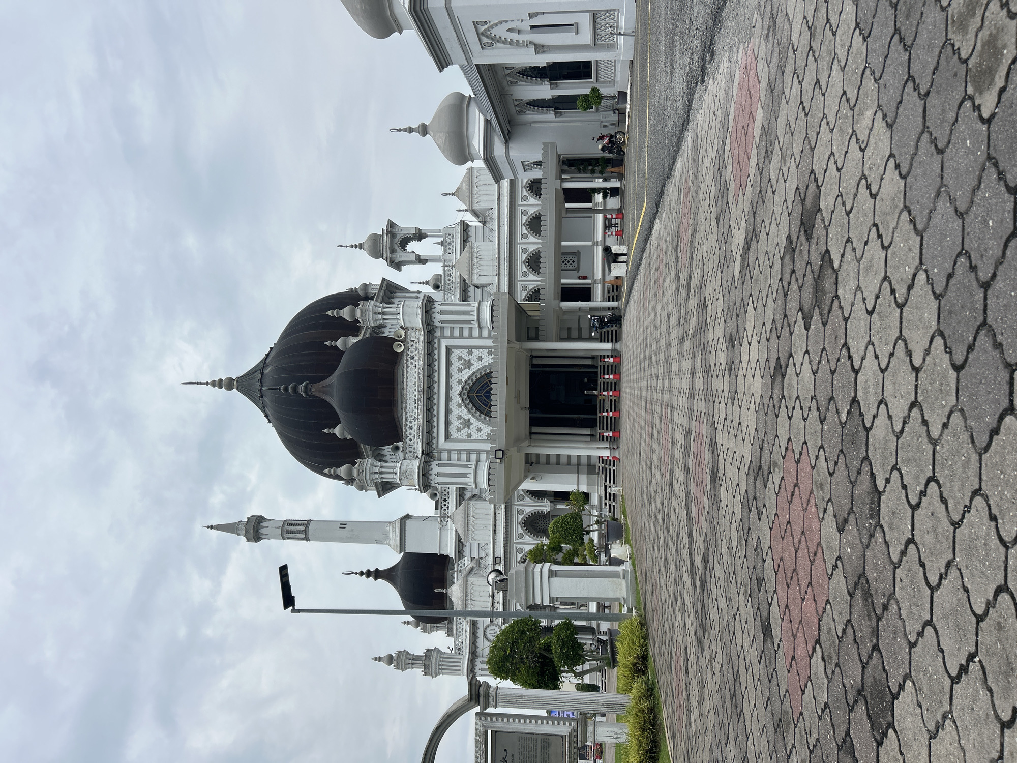 The image captures the grand architecture of the Zahir Mosque, a prominent landmark in Alor Setar, Malaysia. The mosque features a striking white facade with intricate gray detailing and is crowned by several dark domes. A tall, slender minaret stands to the left of the main structure. In front of the mosque, a wide paved walkway composed of interlocking gray and red patterned tiles leads towards the entrance. Several orange traffic cones are placed at intervals along the walkway, and a street lamp is visible on the left. The sky is overcast with gray clouds. Two motorcycles are parked on the right side of the paved area in front of the mosque's extended structures, and a person is seated on one of them. The overall scene suggests a place of worship, likely on a cloudy day, with some activity around the entrance.