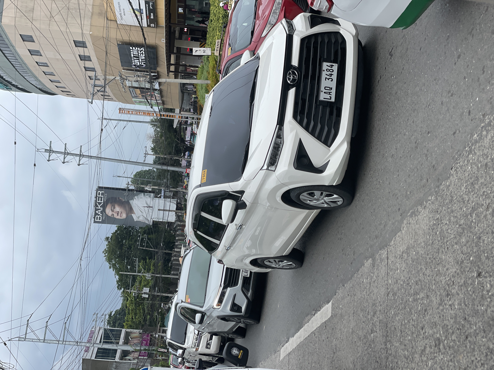 A street scene in Davao City, Philippines, shows a line of cars on a road under an overcast sky. The foreground features a white MPV with a license plate that reads "LAQ 3484". To its left, a white SUV is partially visible, and further back, another white vehicle. In the background, buildings line the street, including a large curved building with multiple stories and signage for "ANYTIME FITNESS" and "BPI". A prominent billboard displays a man's face with the text "BAKER" and "MEDIC AESTHETICS". Overhead, a complex network of electrical wires crisscrosses the sky, supported by utility poles. The street appears to be a busy commercial area.