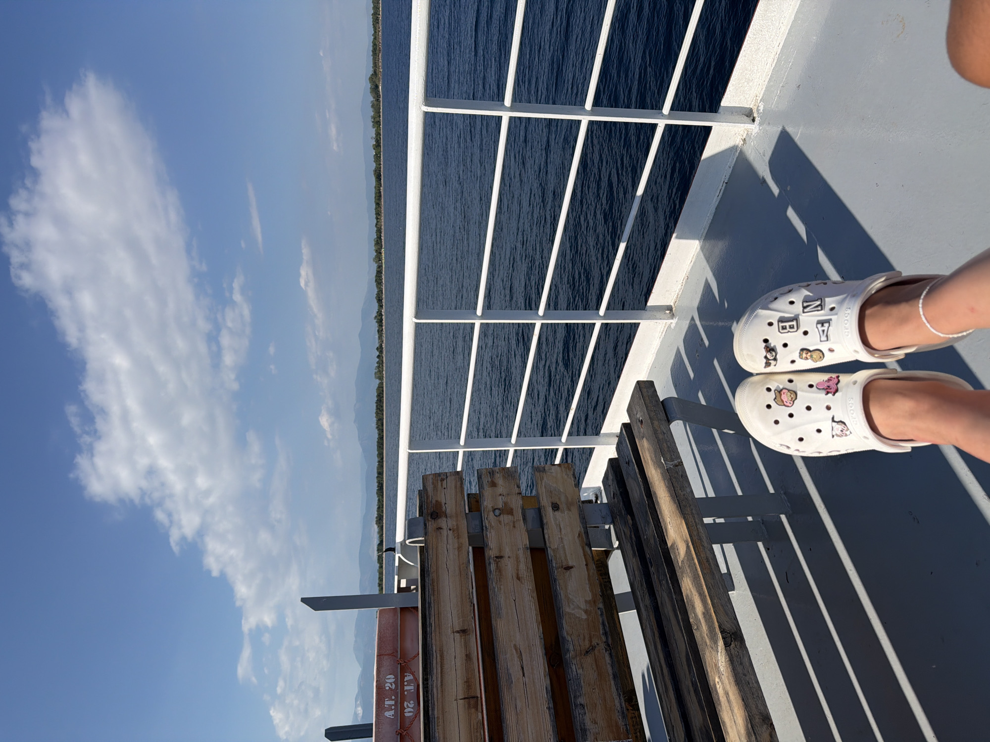 The image captures a view from the deck of a ferry or boat in Keramoti, Greece. In the foreground, a person's legs are visible, wearing white Croc shoes adorned with various colorful charms, including what appear to be cartoon characters and letters. The shoes are positioned on the grey deck of the vessel. To the left, a weathered wooden bench with metal supports is present, casting long shadows across the deck. A life preserver ring, orange with white lettering, is affixed to a railing behind the bench. The middle ground is dominated by the deep blue sea, with white railings of the vessel running parallel to the water. In the background, a distant coastline is visible, featuring a line of trees and rolling hills or mountains under a bright blue sky with scattered white clouds. The overall scene suggests a sunny day at sea, likely a journey to or from Keramoti.