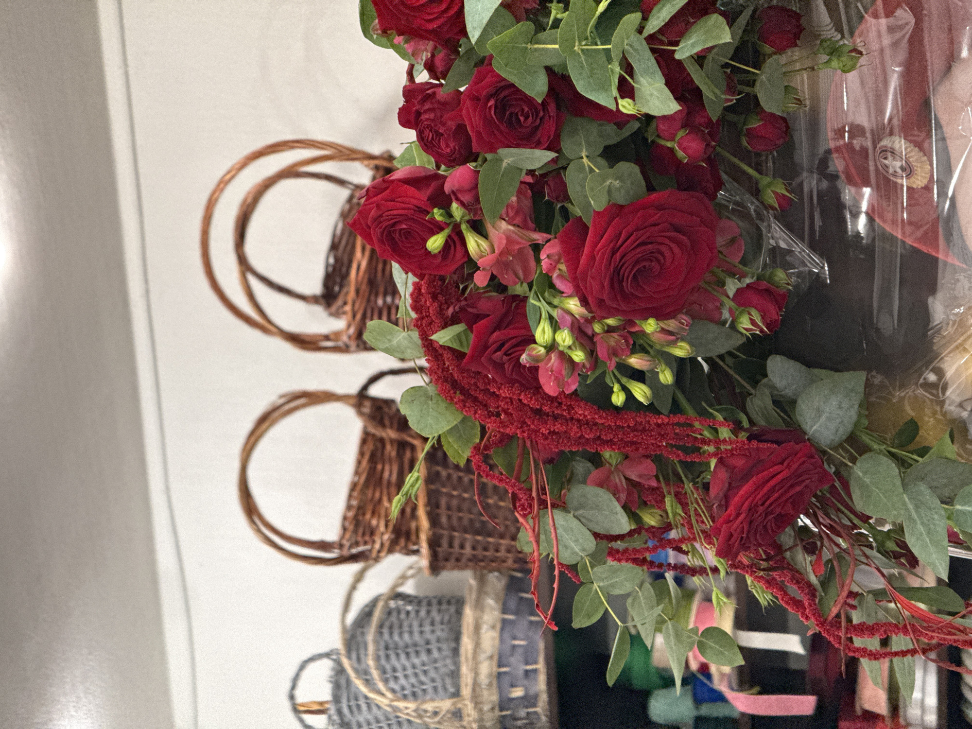 This image captures a close-up view of a vibrant bouquet of red roses and other floral elements, likely arranged by a florist. Behind the flowers, several woven baskets of varying sizes and materials are visible, some stacked on shelves. The baskets appear to be traditional in style, some made of wicker and others of a gray, textured material. In the background, shelves are stocked with various items, including what appear to be spools of ribbon or craft supplies in different colors, suggesting a retail environment such as a flower shop or craft store. The overall impression is one of abundance and natural beauty, possibly in preparation for an event or sale. The scene is indoors, with soft lighting that highlights the rich red hues of the flowers. The specific location in Vladikavkaz, Russia cannot be determined from the visual information provided.