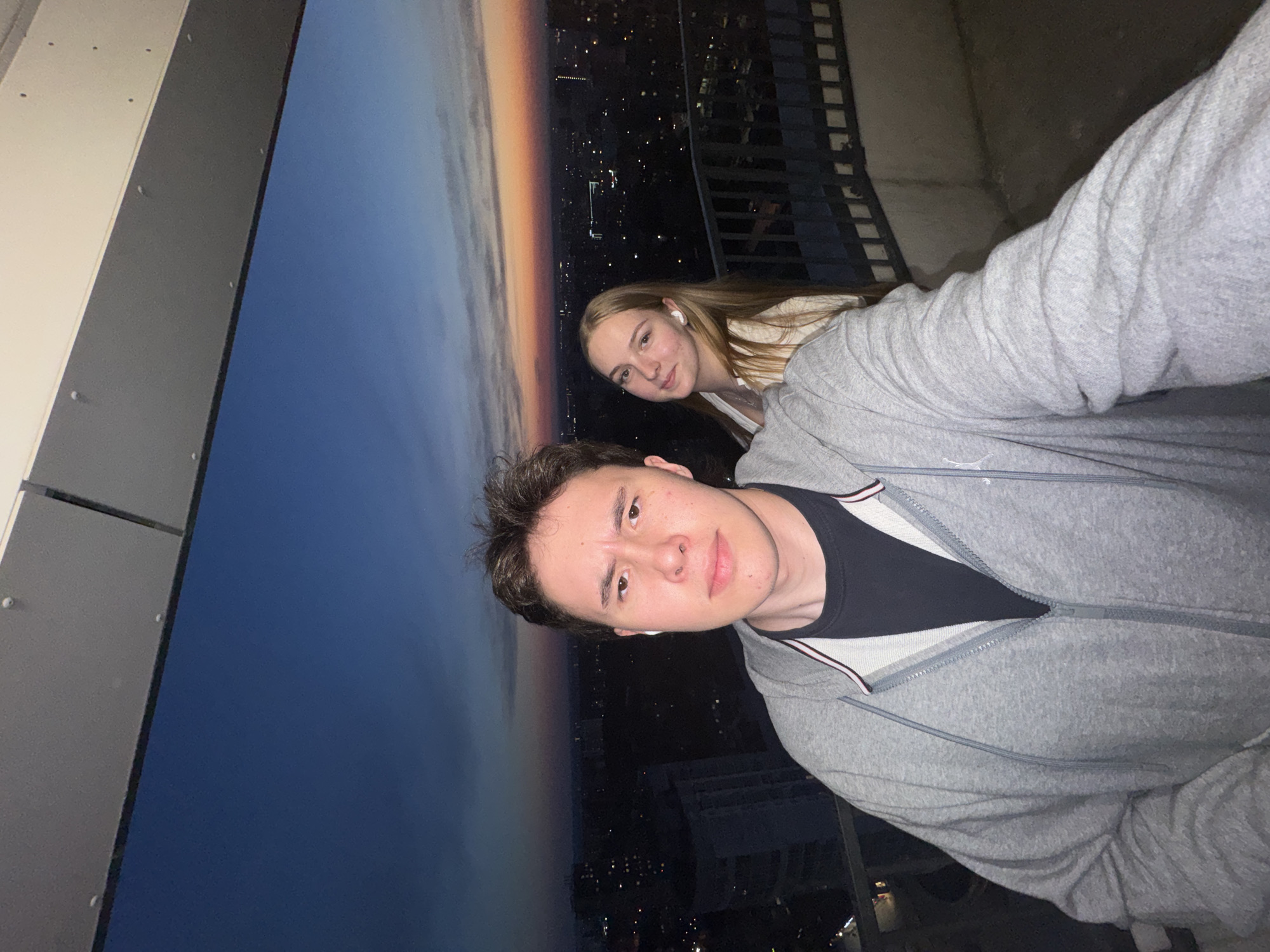 Two people, a man and a woman, are posing for a selfie. The background appears to be a cityscape at dusk, with a visible skyline and city lights. The man is closer to the camera, wearing a gray zip-up hoodie over a dark-colored shirt. The woman stands behind him, wearing white earbuds. The photo is taken from an elevated position, possibly on a balcony or rooftop, with a wall or structure partially visible on the left side of the frame. The location is likely Perm, Russia, based on the urban landscape.