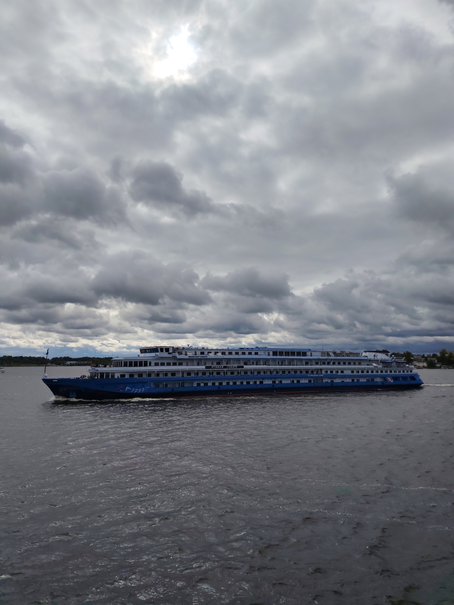 In the city of Kalyazin, Russia, a large blue and white multi-deck river cruise ship, identified by the lettering "ЛУННАЯ СОНАТА" (Moonlight Sonata) on its side, sails on a body of water under a heavily clouded sky. The ship is positioned in the middle ground, moving from left to right. Its hull is blue with a red waterline, and it features multiple rows of windows on its upper decks, which are white. The sky is filled with dramatic, dark grey clouds, with a brighter area indicating the sun is obscured. The water is choppy, reflecting the overcast conditions. The shoreline is visible in the distance on the right, with some trees and buildings, and on the left, a bridge can be seen. There are no people clearly visible on the deck of the ship.