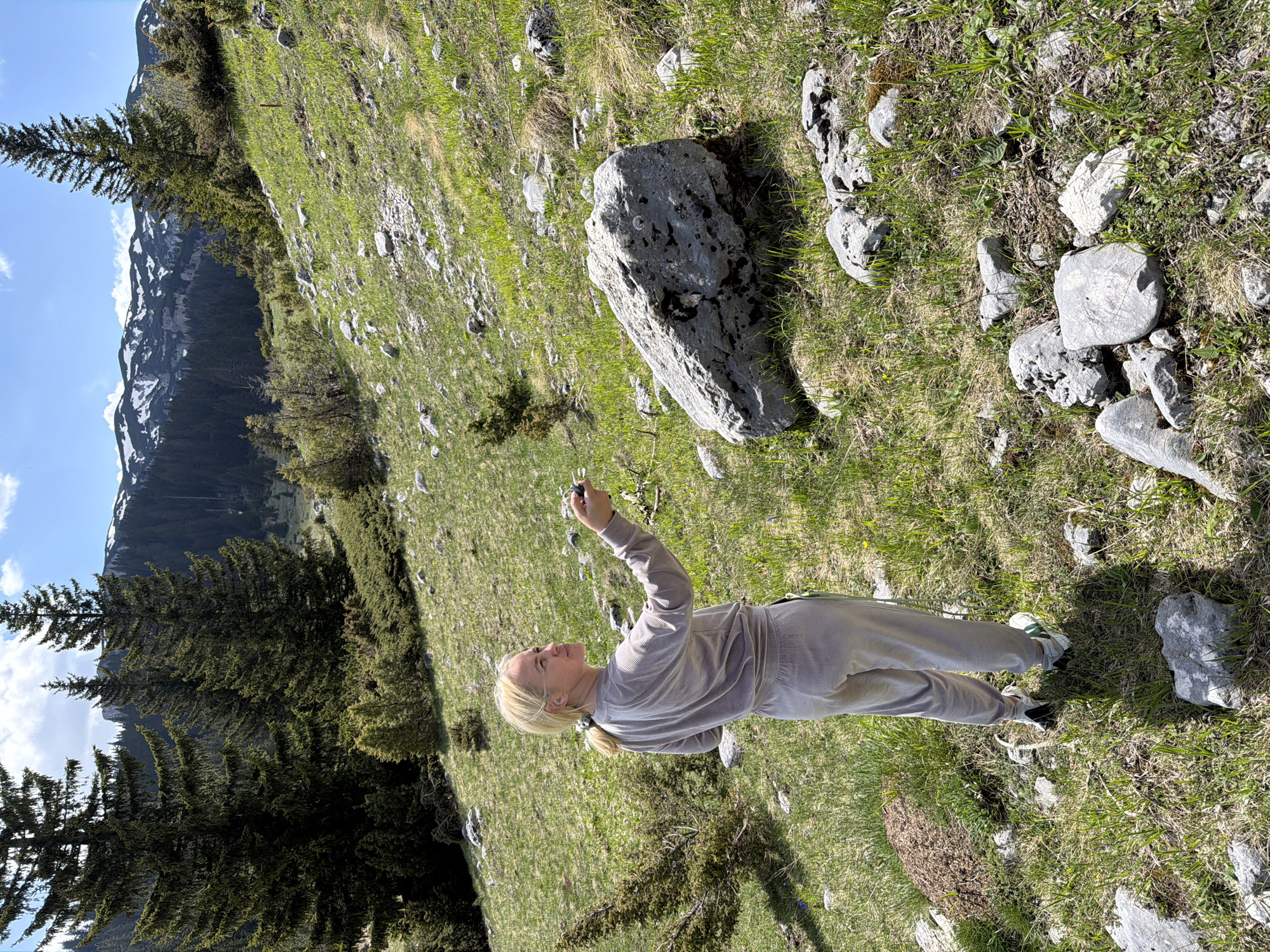 A woman stands on a grassy, rocky hillside in Bosača, Montenegro. She wears a light-colored pantsuit and sneakers and is holding a small object above her head. The hillside is covered in grass and rocks, with a large boulder near her. In the background, there are evergreen trees and a mountain with patches of snow. The sky is partly cloudy.
