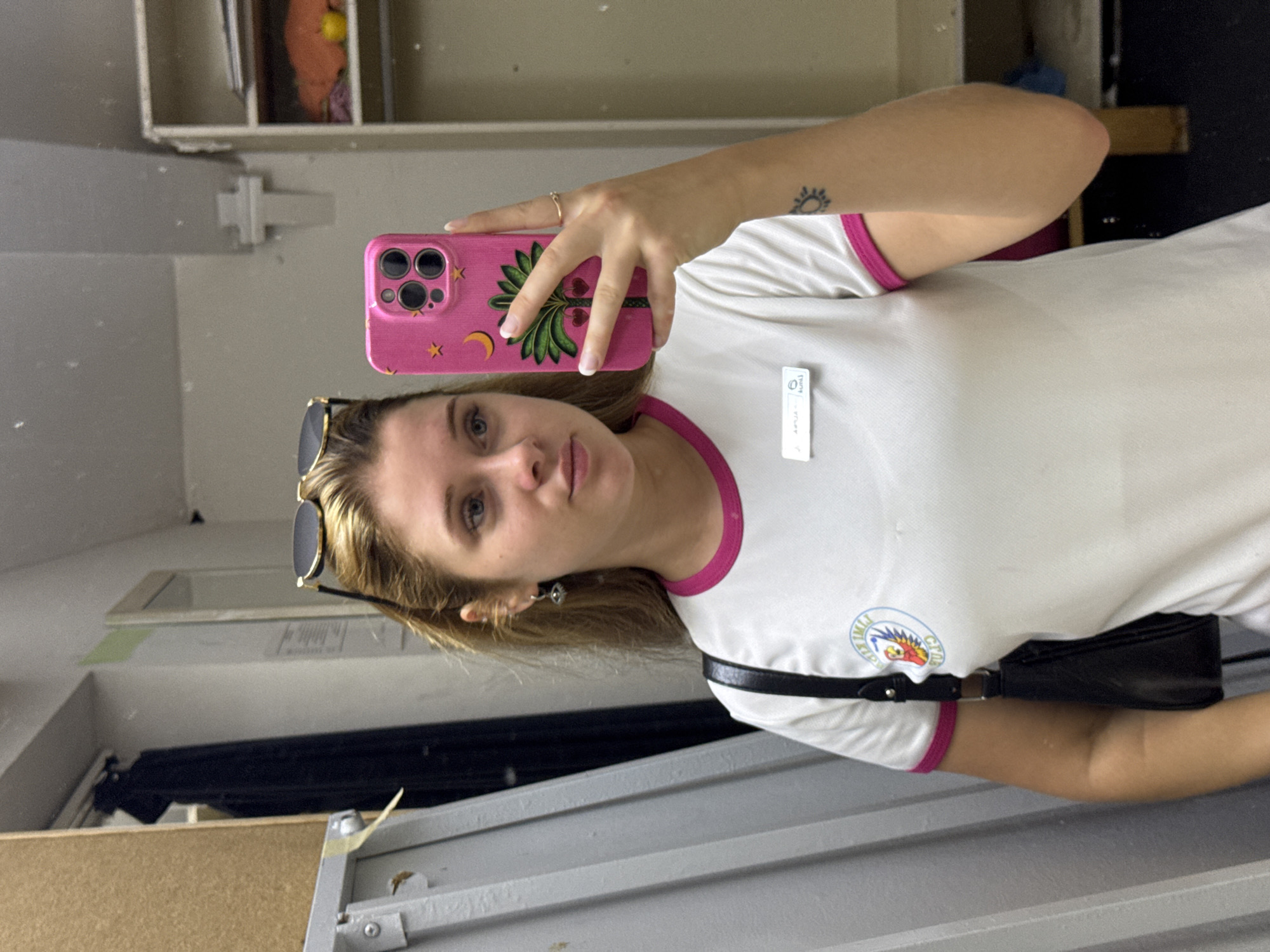 A young woman with blonde hair and blue eyes is taking a mirror selfie in what appears to be a changing room or a storage area. She is wearing a white t-shirt with pink trim and a logo on the left chest. She has a black purse slung over her shoulder and sunglasses perched on her head. She is holding a pink phone with a palm tree design. There is a name tag on her t-shirt. A tattoo of a sun is visible on her right arm. The background shows shelves, clothing racks, and a grey corrugated wall. There is no visible indication of the location being Aksu, Turkey.