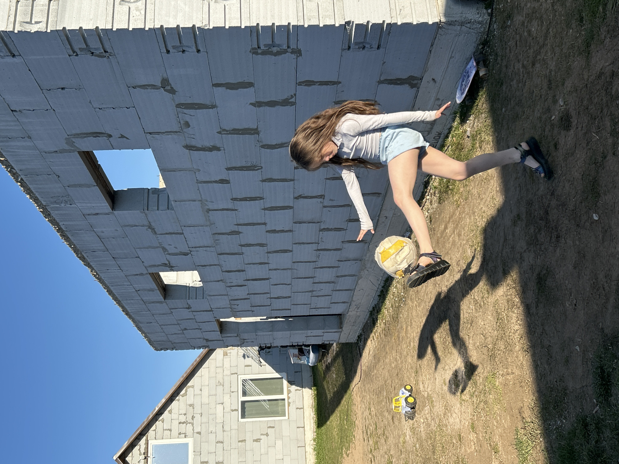 A girl is jumping toward a soccer ball in the city of Chaikovsky, Russia. The background includes an unfinished concrete building and a blue sky. The ground is a mix of dirt and grass, with children's toys scattered around.