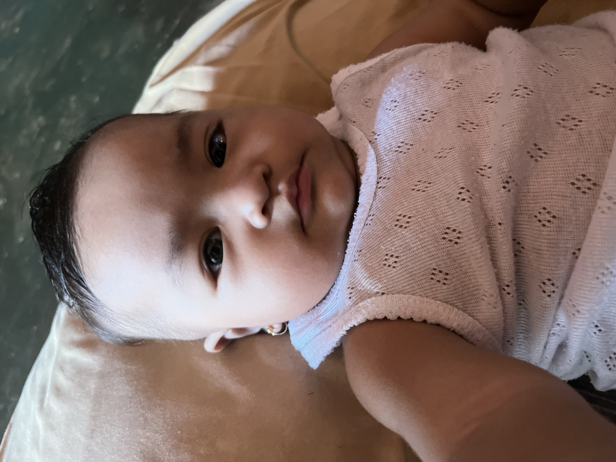 A close-up, high-angle selfie shows a baby lying down, looking directly at the camera. The baby is wearing a white, sleeveless top with a lace-like pattern and a small gold hoop earring in its left ear. Its dark hair is styled upwards. The background is out of focus, but appears to be a soft, possibly cushioned surface, possibly a bed or couch, with a warm, light brown hue. The lighting suggests an indoor environment. The scene is likely from Kruengmate, Indonesia, given the provided context.