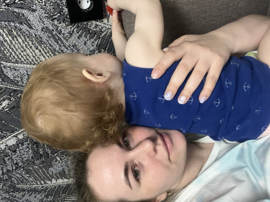 An adult female and an infant are present in what appears to be an indoor setting in Chaikovsky, Russia. The female, positioned on the left, has light skin, green or hazel eyes, and light brown hair partially visible. She is wearing a light-colored garment, possibly white with a blue-green pattern. Her left hand, with white nails featuring small colorful speckles, supports the infant. The infant, facing away from the camera towards the right, has light skin and reddish-blonde curly hair, and is dressed in a blue sleeveless onesie adorned with white anchor patterns. The infant's right hand holds a small red object. In the upper right background, a black rectangular object containing a circular silver or white component is visible against a dark gray and white patterned fabric, possibly upholstery or wallpaper, featuring an abstract or leaf-like design. The subjects appear to be supported by a brown, upholstered surface, likely a sofa or armchair.