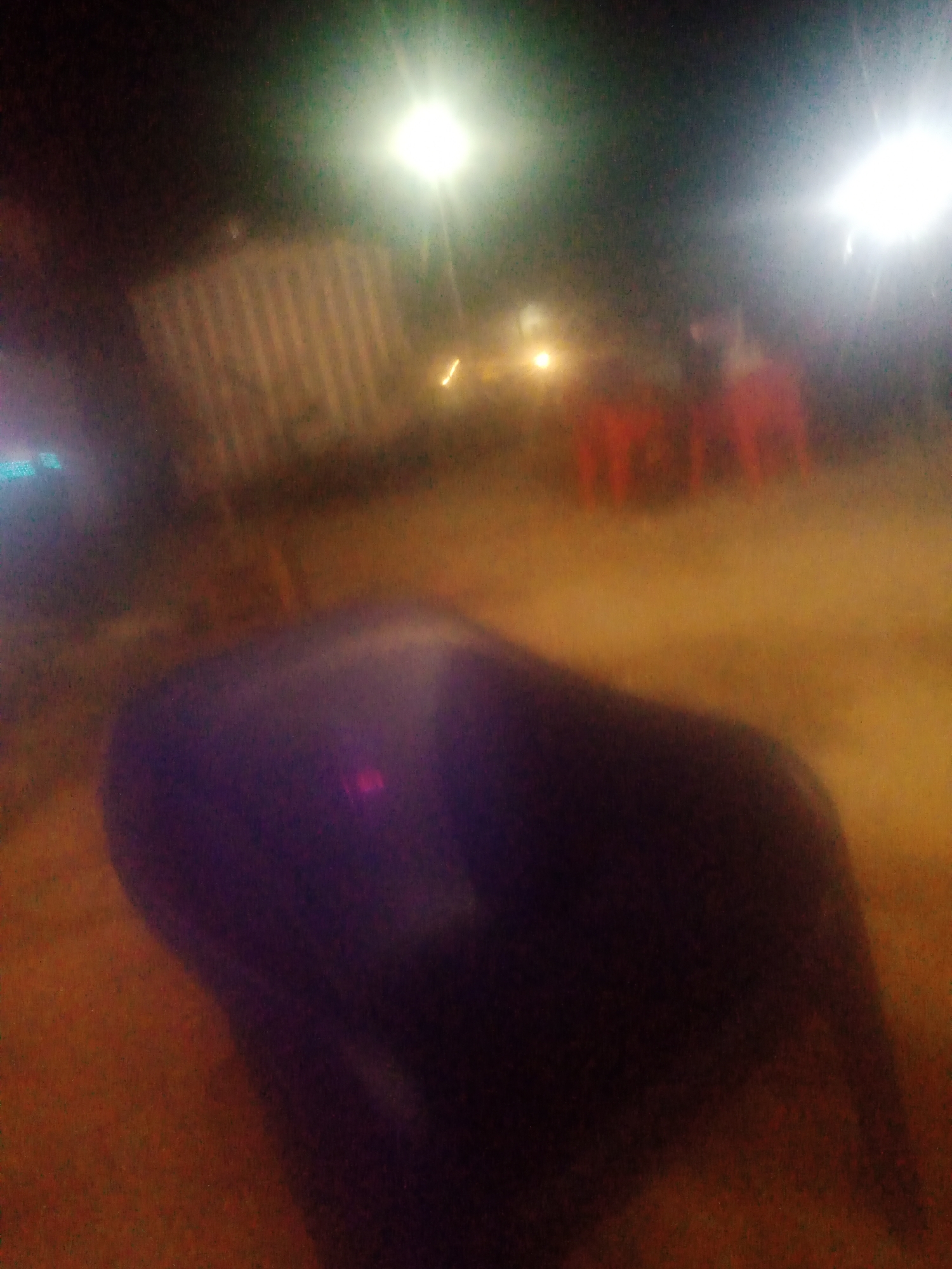The image depicts a dimly lit outdoor scene, likely at night, with prominent artificial lighting casting bright glows and lens flares. In the foreground, a large, dark, and out-of-focus object dominates the lower portion of the frame, appearing to be a piece of furniture or perhaps a person's back. In the mid-ground, several red chairs are visible, suggesting a seating area. Behind the chairs, a structure with corrugated metal siding is discernible. The overall environment appears to be dusty or sandy, with a warm, earthy tone. The lighting suggests an urban or semi-urban setting, possibly a roadside stall or an informal gathering place. Without further context, it is not possible to definitively pinpoint the location within Jalingo, Nigeria, but the elements present are consistent with common informal business or social spaces found in such environments.