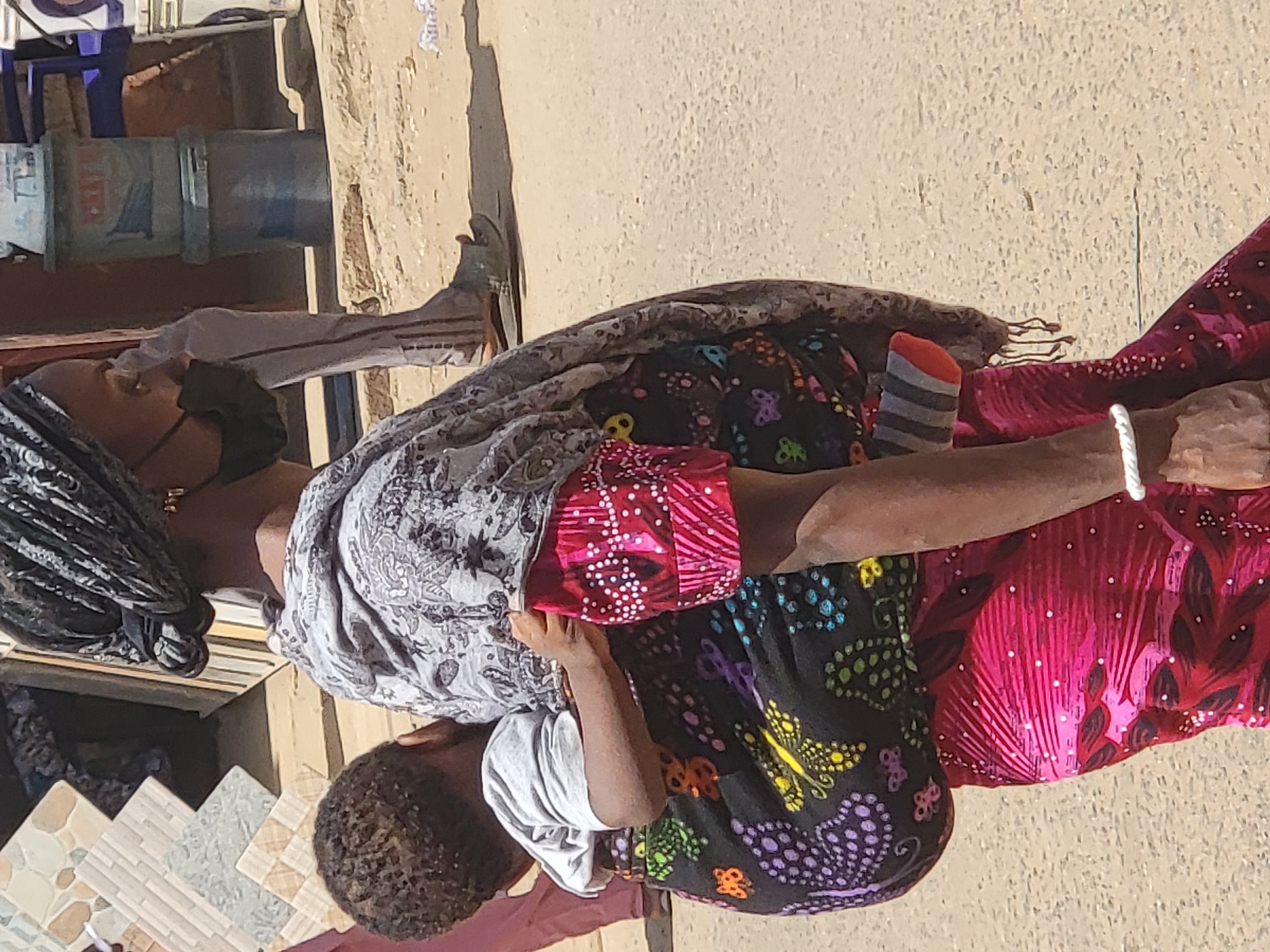 Two people are sitting on the ground in an urban environment, possibly Maiburiji, Nigeria. One person wears a face mask and a patterned head covering, while the other has short, curly hair and is wearing a white shirt. The person closest to the camera is wearing brightly colored patterned clothing, including a black and multicolored skirt and a pink patterned top. One of their feet is covered with a gray and orange striped sock. A white rope or bracelet is around their wrist. They are seated on a surface comprised of multi-colored tiles with a beige background. In the background are sections of a building, including a section with what appears to be faded and peeling paint, along with a section that might be part of a blue chair or water pipe.