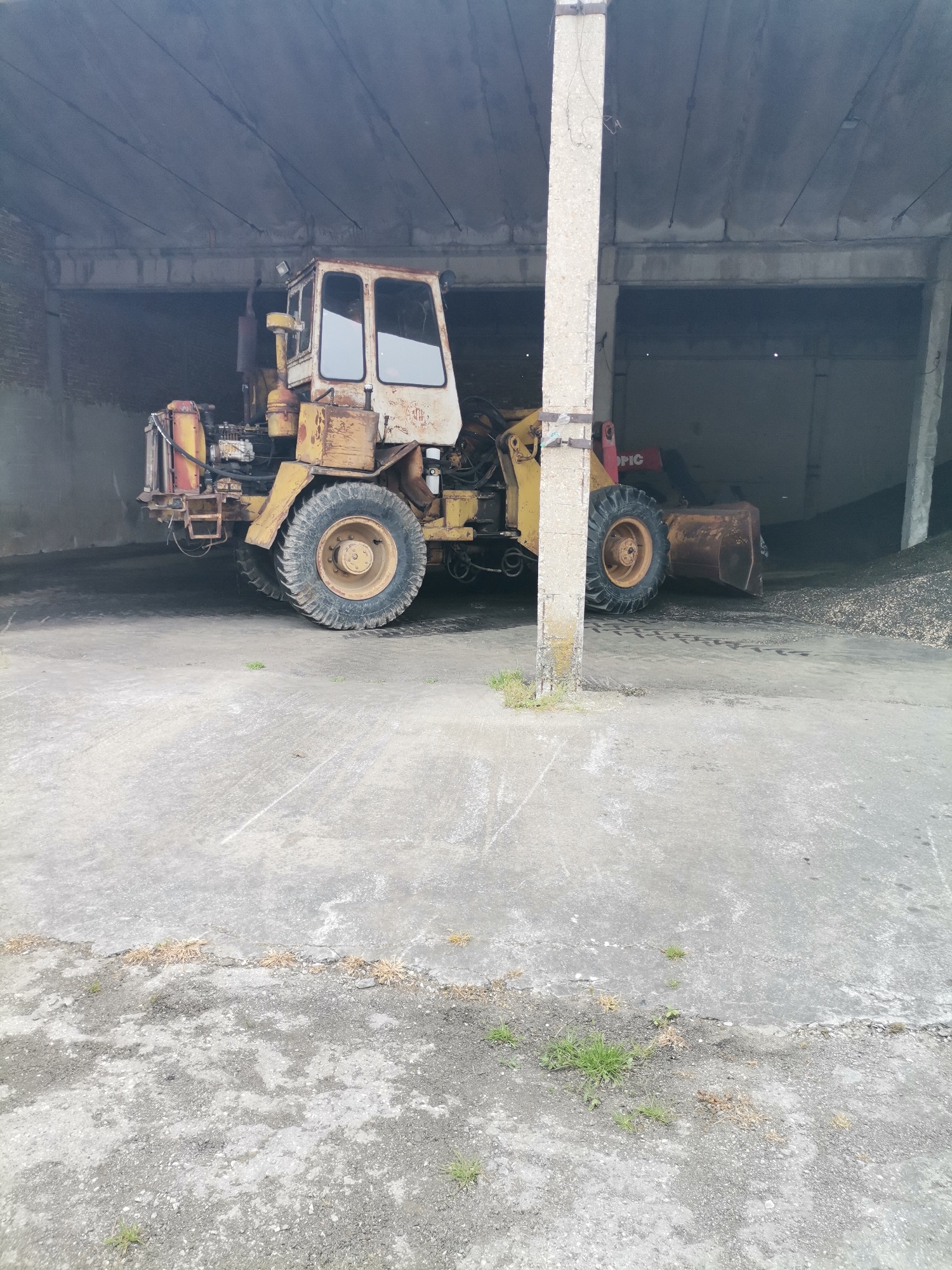An old, weathered yellow and orange front-end loader is parked inside a large, open-sided concrete structure, likely a storage shed or industrial building, in Kochovo, Bulgaria. The image is taken from a slightly low angle, looking towards the back of the shed.
The loader, the primary subject, shows significant signs of rust and wear, with chipped paint and exposed mechanics. It has a large cabin with glass windows, prominent, deeply-treaded tires, and a heavy-duty bucket resting on the ground to its right. Visible details on the loader include various hoses, metal components, and what appears to be an exhaust pipe. A faint, weathered logo is present on the side of the cabin. On the red hydraulic arm component, to the right of the central pillar, the letters "OPIC" are visible, suggesting part of a brand name.
The structure features a concrete floor, concrete pillars, and a concrete slab roof with visible reinforcement lines. A brick wall is visible in the dim background, to the left of the loader. Behind the loader's bucket, a large pile of dark, granular material, possibly gravel, seeds, or coal, is visible on the ground.
The lighting is diffused and dim, consistent with an indoor setting, suggesting it is daytime but shielded from direct sunlight. The concrete ground in the foreground is cracked and dirty, with small patches of green and dry grass growing through the fissures. The overall scene conveys an impression of age, utility, and possibly disuse or maintenance.