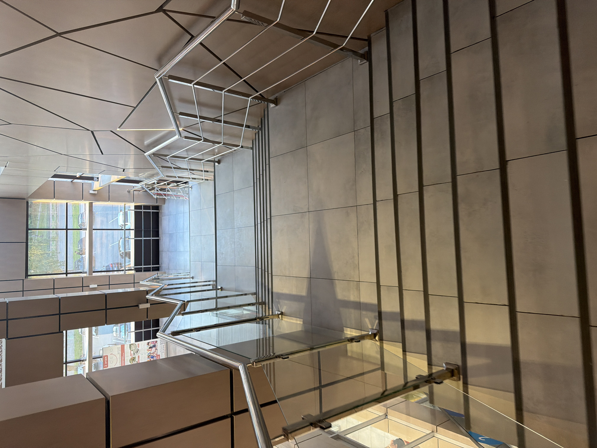 An indoor staircase with tiled steps and modern metal railings with glass panels leads down. The walls are paneled with a geometric pattern. Large windows on the left reveal an outdoor scene with parked cars and trees. The lighting suggests an interior space. There are no people clearly visible in the image.