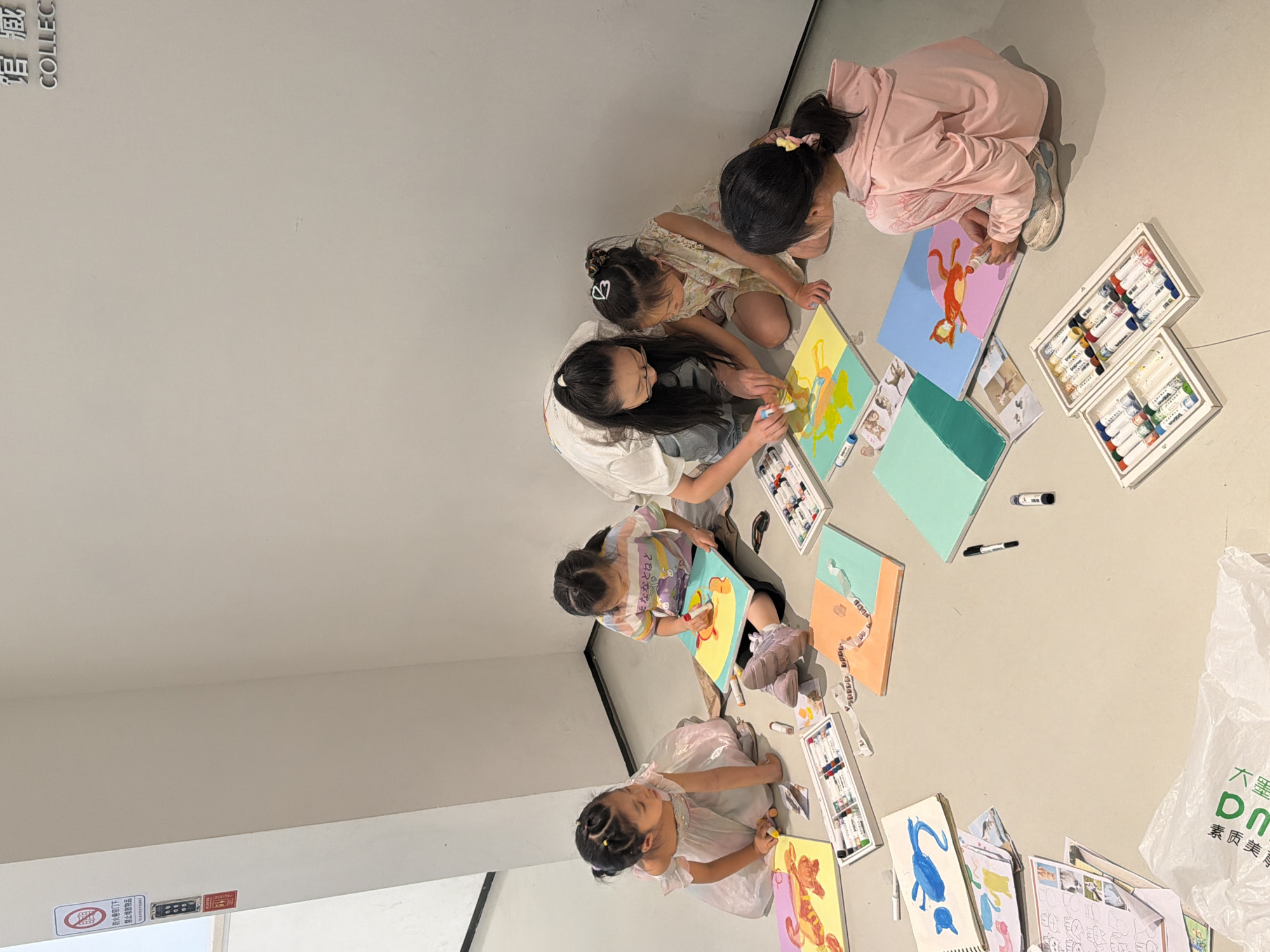 This image captures an indoor art activity taking place in the Chongqing High-tech Zone, China. One adult woman and five young girls are sitting on a light-colored floor, engaged in painting on small canvases.
The adult woman, positioned centrally, is wearing a white short-sleeved shirt, light-colored jeans, and glasses, with her dark hair tied in a high ponytail. She is kneeling and leaning forward, actively demonstrating or assisting with painting on a yellow and teal canvas using a white marker-like paint stick. Three girls are immediately around her.
To her left, a girl in a light pink iridescent dress with her hair in two small ponytails sits, holding a white paint stick and observing a yellow canvas featuring a reddish-orange mythical creature. Next to her, a girl in a striped t-shirt and light-colored pants, with her hair in two ponytails, is focused on painting a red-orange figure on a yellow and teal canvas. To the adult's right, a girl in a patterned light-colored dress with white hair clips sits, observing the adult's actions.
Further to the right, a fifth girl in a pink long-sleeved top and light-colored sneakers is actively painting a red-orange cat-like figure on a light blue and pink canvas. Her dark hair is tied in a high ponytail with a yellow elastic. A sixth girl's lower body is partially visible to the upper right of this girl.
The floor is a uniform light gray or beige. Scattered around the participants are numerous art supplies, including multiple white plastic trays filled with colored paint sticks or markers, loose black markers, and various small tubes or containers. Several completed or partially completed canvases with solid colored backgrounds (yellow, teal, light blue, pink, orange) and painted figures are visible. A stack of paper with drawings and sketches is on the floor to the left, along with a white plastic bag bearing green Chinese characters in the bottom-left corner.
The background consists of plain white or off-white walls. On the left wall, a white sign with a red circular 'no' symbol and Chinese text warns against obstructing fire doors, below which are a red control panel with black buttons and a small red sticker. On the top-right wall, a silver metallic sign displays the Chinese characters "馆 藏" and the partial English word "COLLEC," indicating a museum or collection setting.