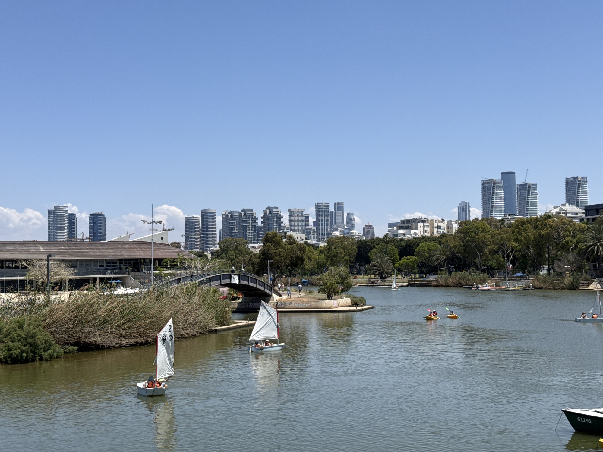 The image shows a tranquil scene in Tel Aviv, Israel, likely taken at Park Hayarkon. The foreground features a body of water, possibly the Yarkon River, with several small sailboats navigating its surface. Two kayaks, each with two occupants, are visible further into the scene. The presence of buildings and trees lines the riverbank.
In the background, a cityscape is visible, revealing modern high-rise buildings that characterize Tel Aviv's urban skyline. A pedestrian bridge is crossing over the water. There are a few pedestrians on the bridge. The sky is clear and blue.
